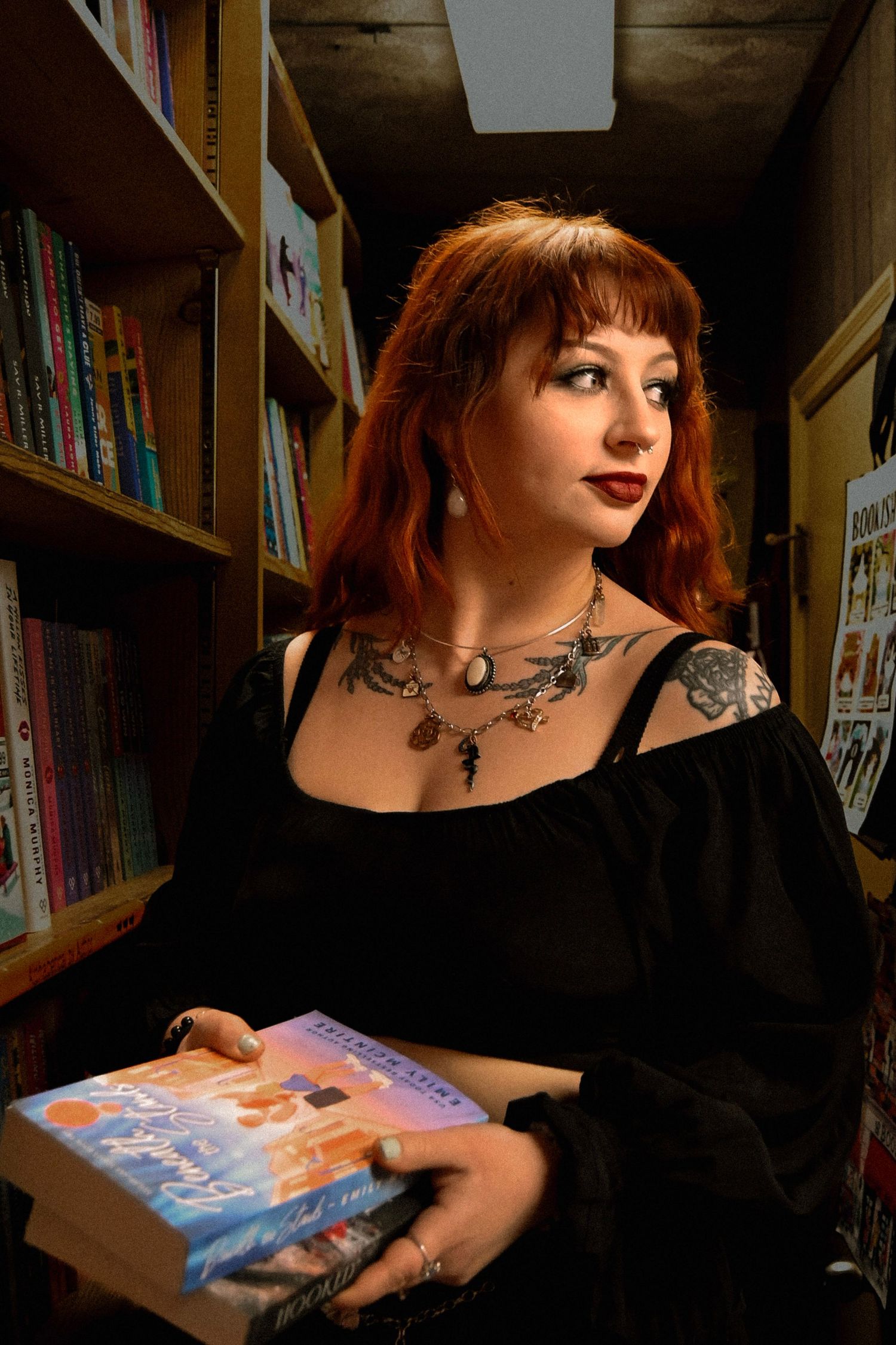 A person with red hair and tattoos browses books in a dimly lit library setting.