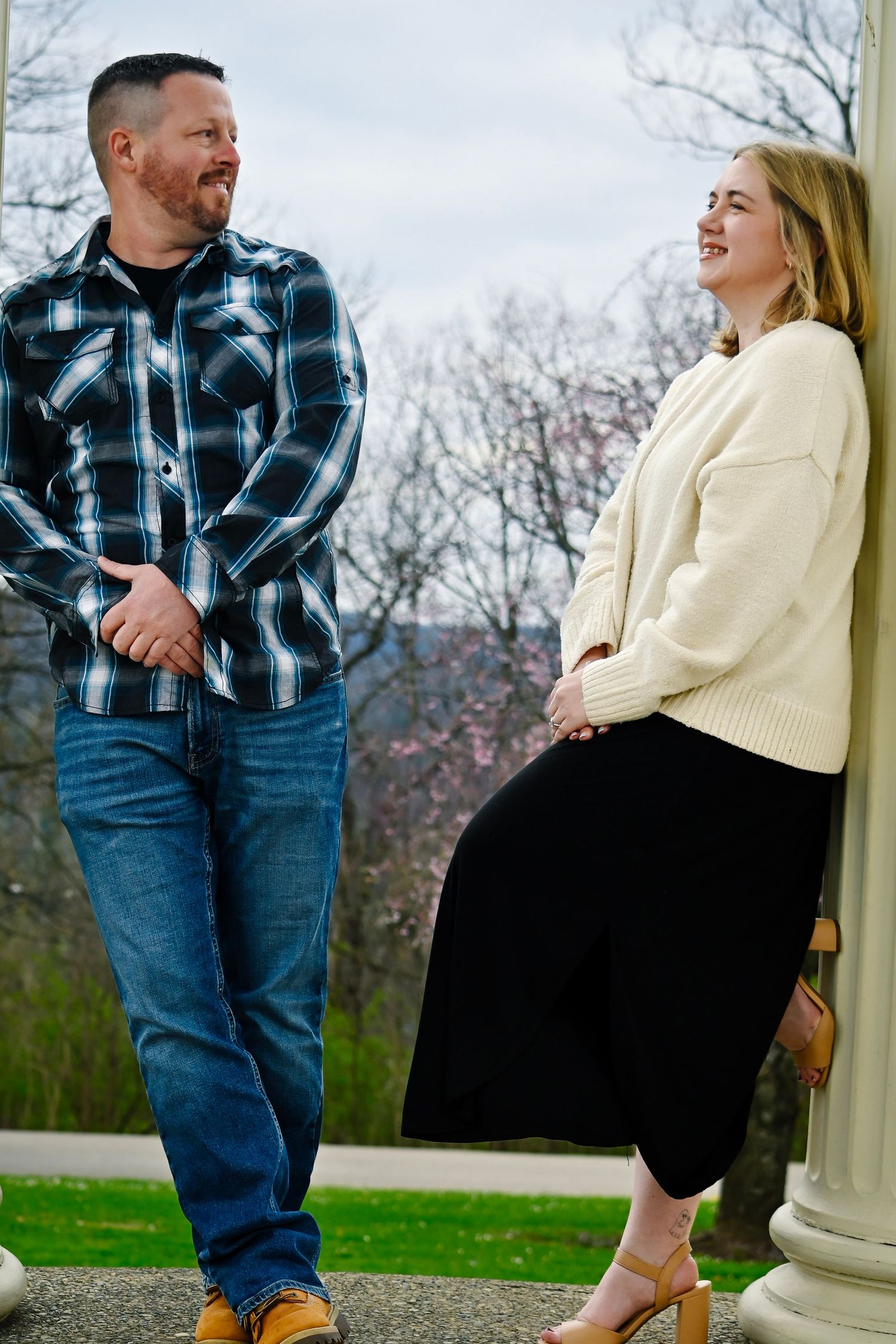 A smiling couple stands by a white column outdoors, man in plaid shirt and jeans, woman in cream sweater and black skirt.