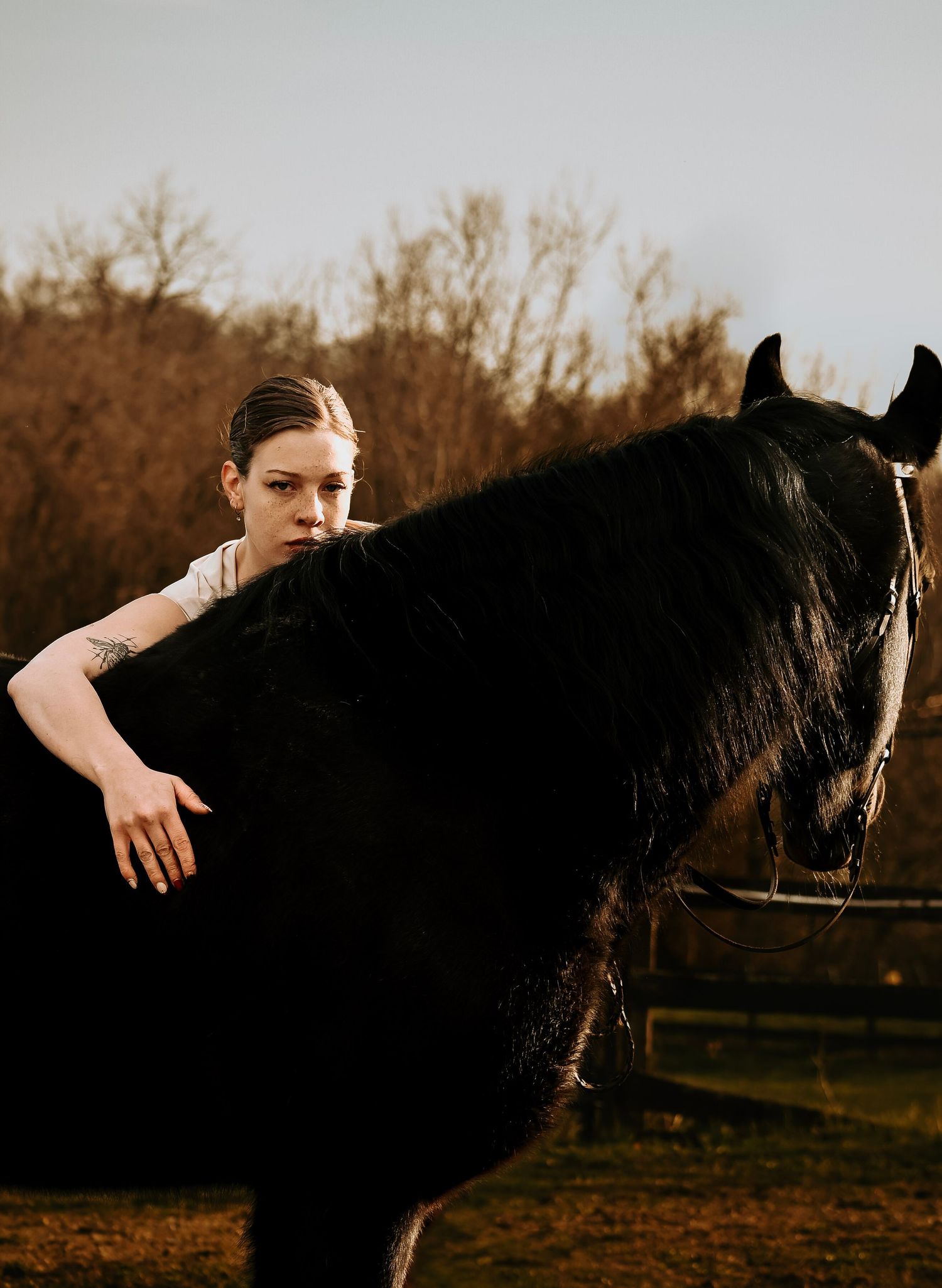 A young woman with a tattoo leans closely against a black horse outdoors, with bare trees in the background.