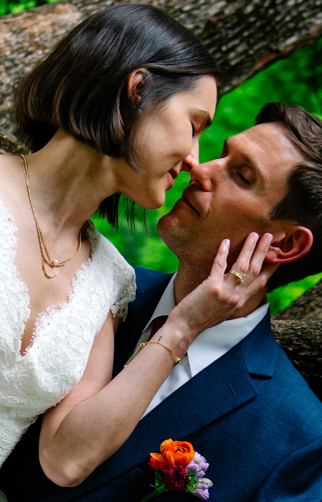 Bride and groom share a romantic kiss outdoors, bride in white lace dress with gold necklace, groom in navy suit with floral boutonniere.