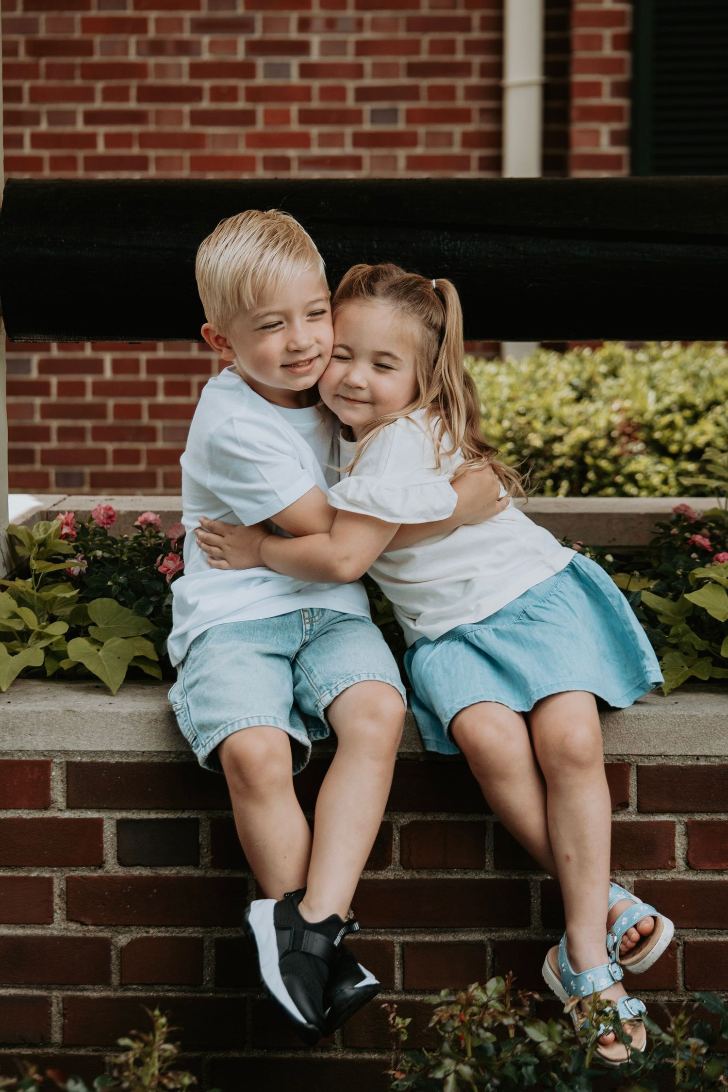 Two young siblings in matching white tees and blue shorts share a sweet hug while sitting on a brick ledge outdoors.