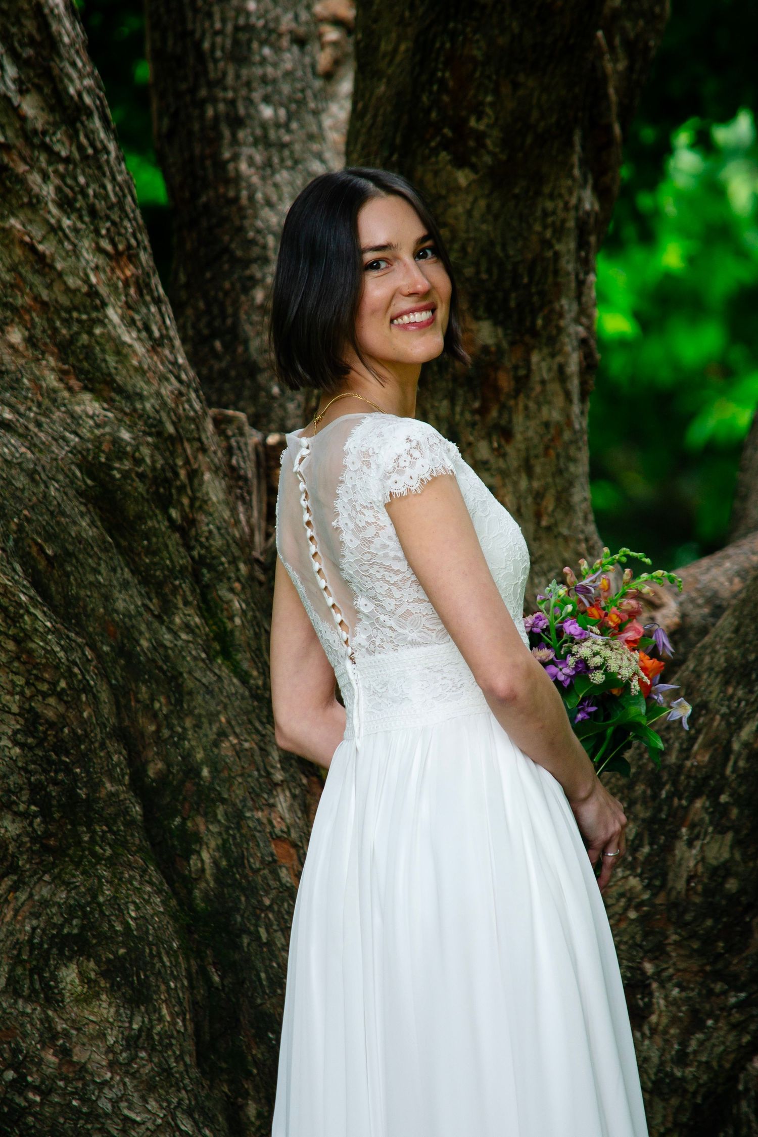 Smiling bride in white lace wedding dress holding colorful bouquet, posing beside large tree trunk in lush green forest.