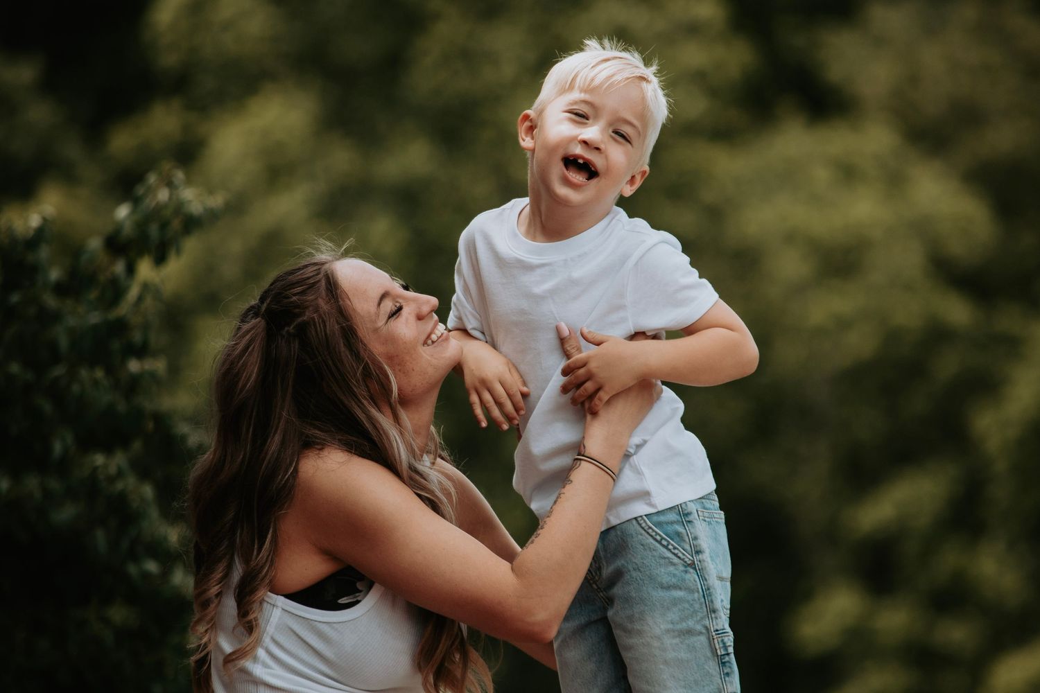 A joyful mother lifts her laughing blonde toddler boy outdoors, both smiling with green trees in the background.