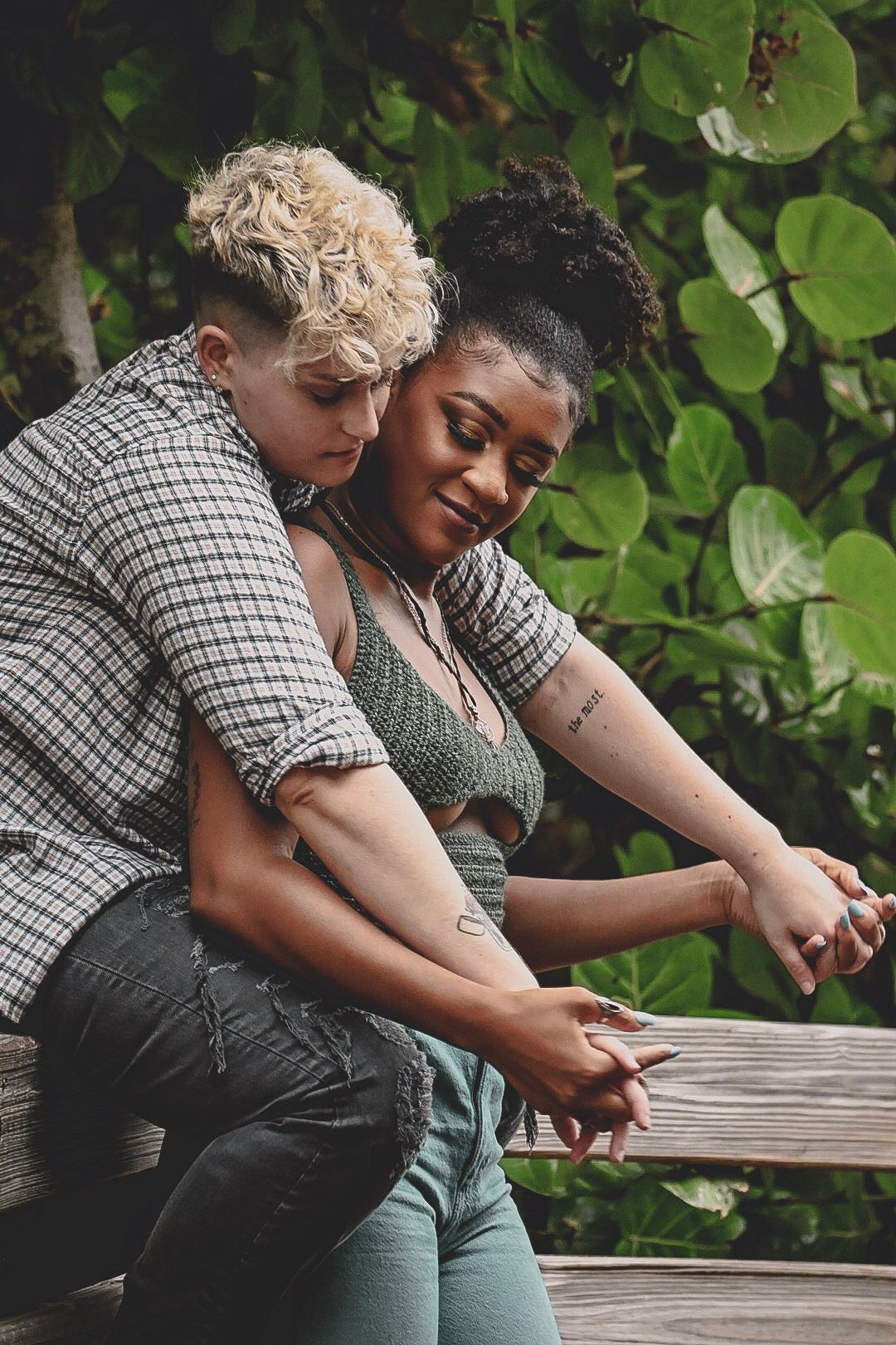 A couple sits close together on a wooden bench surrounded by lush green foliage on a sunny day.