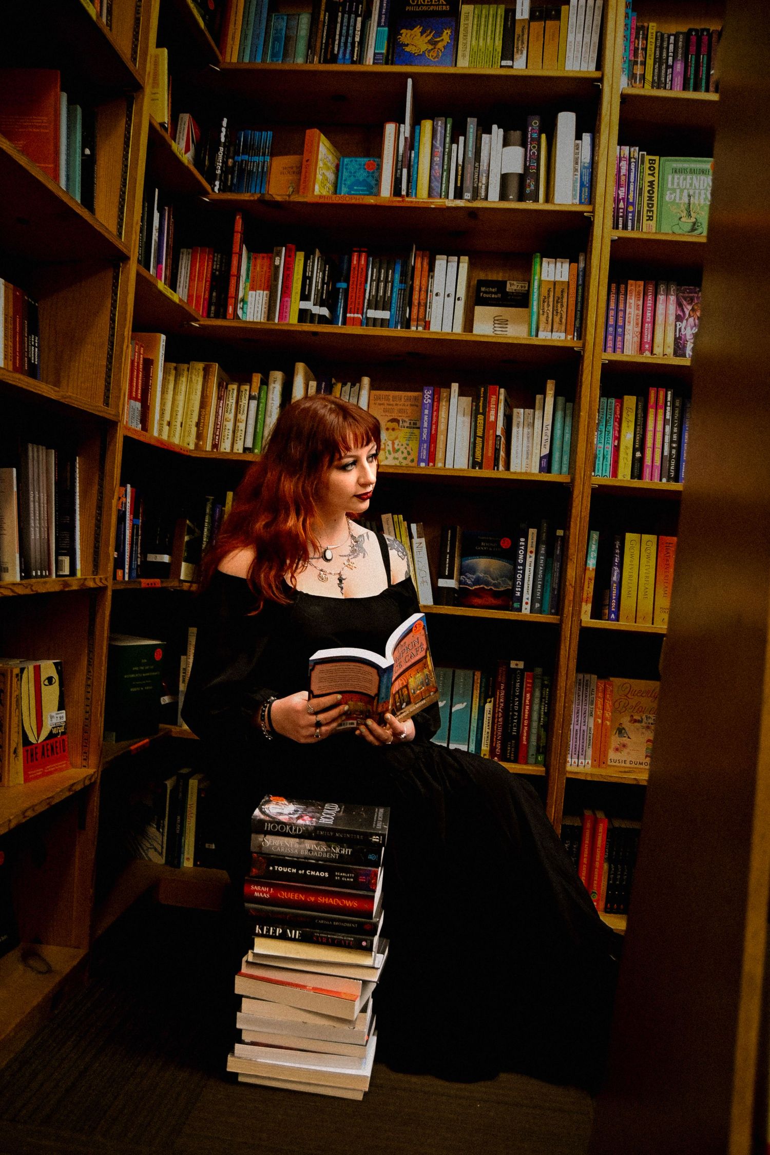 A reader in a black dress sits among library shelves with a stack of books beside them in warm lighting.