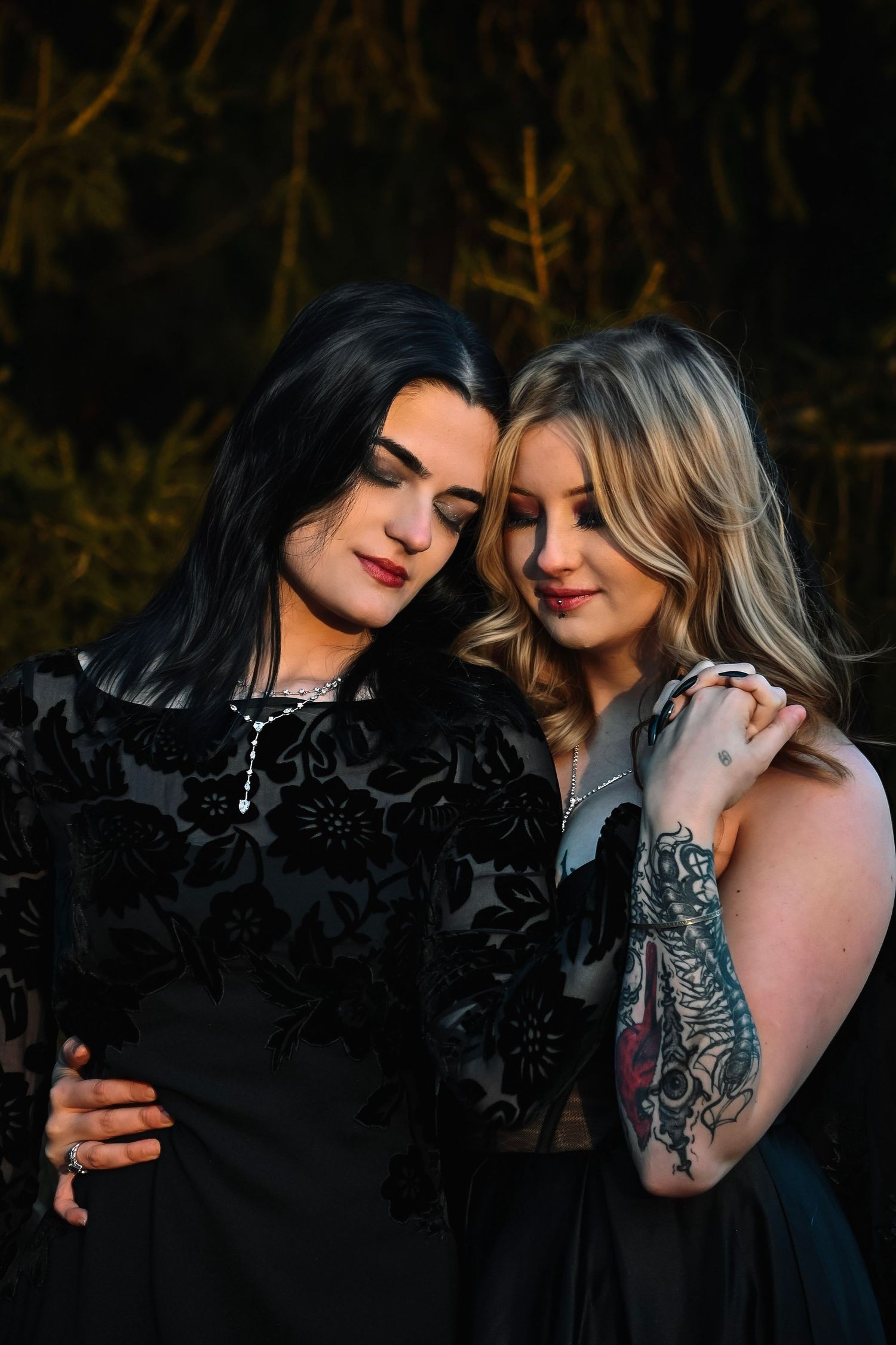 Two women in black dresses pose together outdoors at dusk, one with dark hair and one with blonde hair.