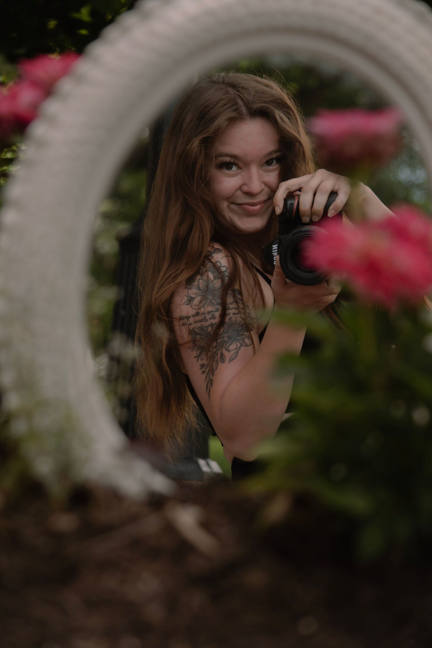 Young woman with tattoos holds a camera in a lush garden framed by a white tire planter with pink flowers.