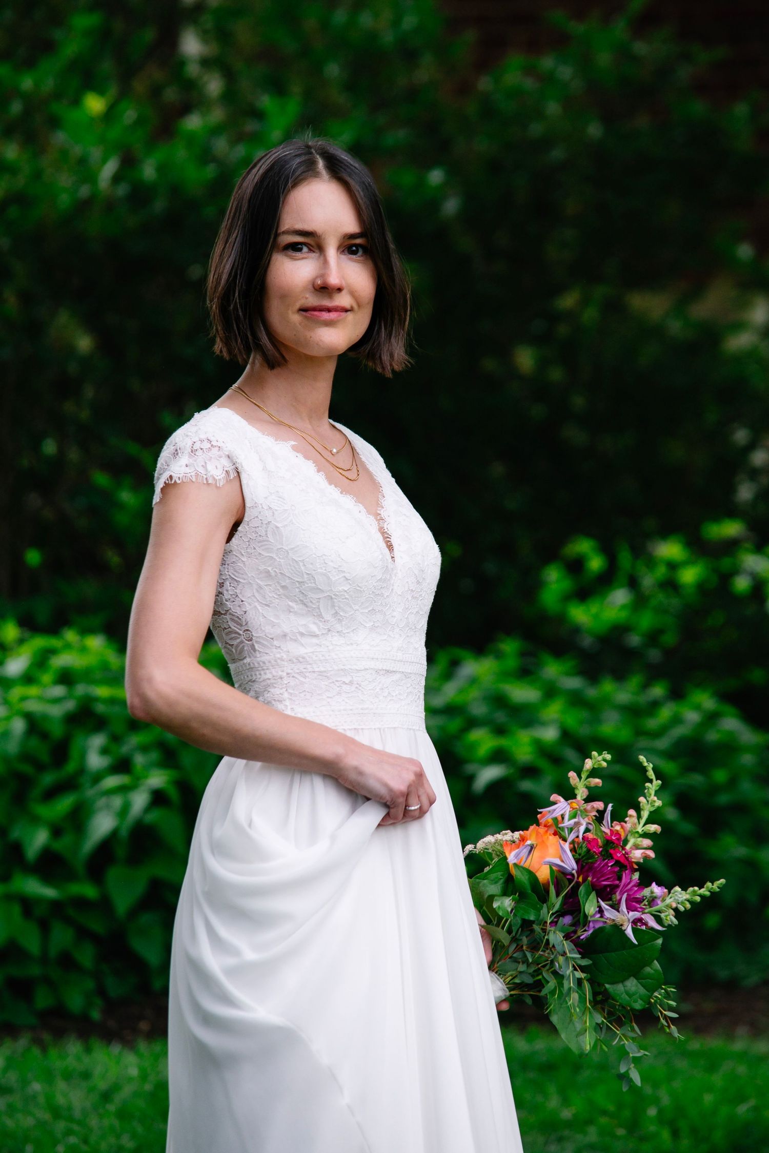 Bride in white lace wedding dress holding colorful bouquet, posing outdoors in lush green garden setting.