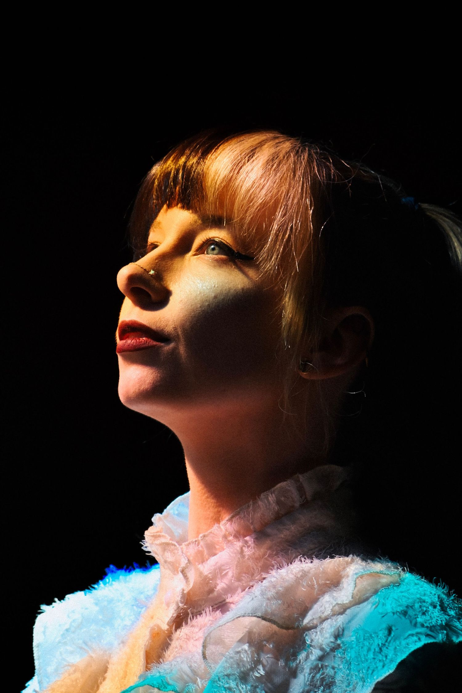 A woman with red hair gazes upward, dramatically lit against a black background, wearing a fluffy teal and white costume.