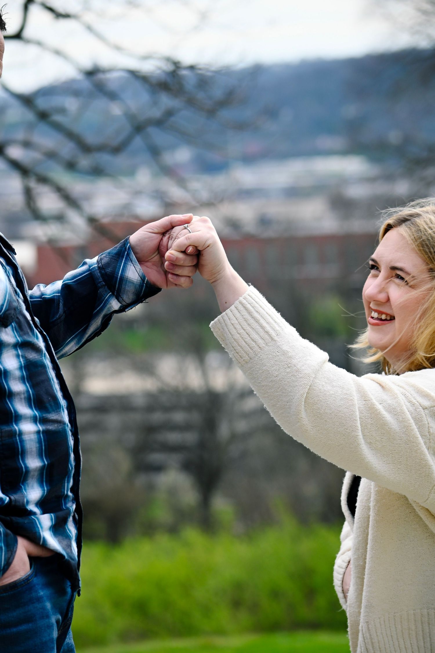 A smiling blonde woman in a white sweater holds hands with a man in a plaid shirt outdoors in a park.