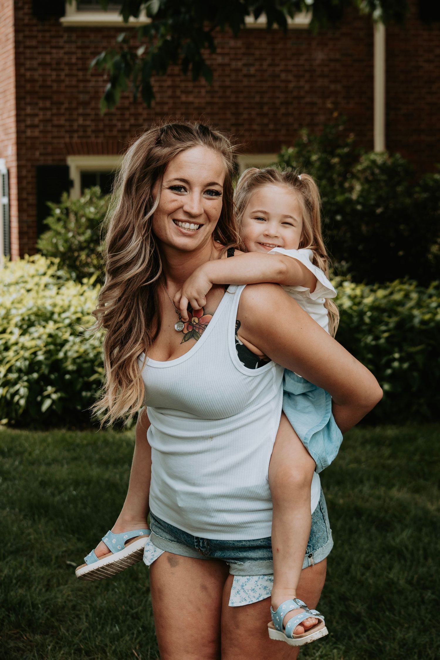 Smiling woman with wavy hair holding a happy little girl on her hip in a green yard.