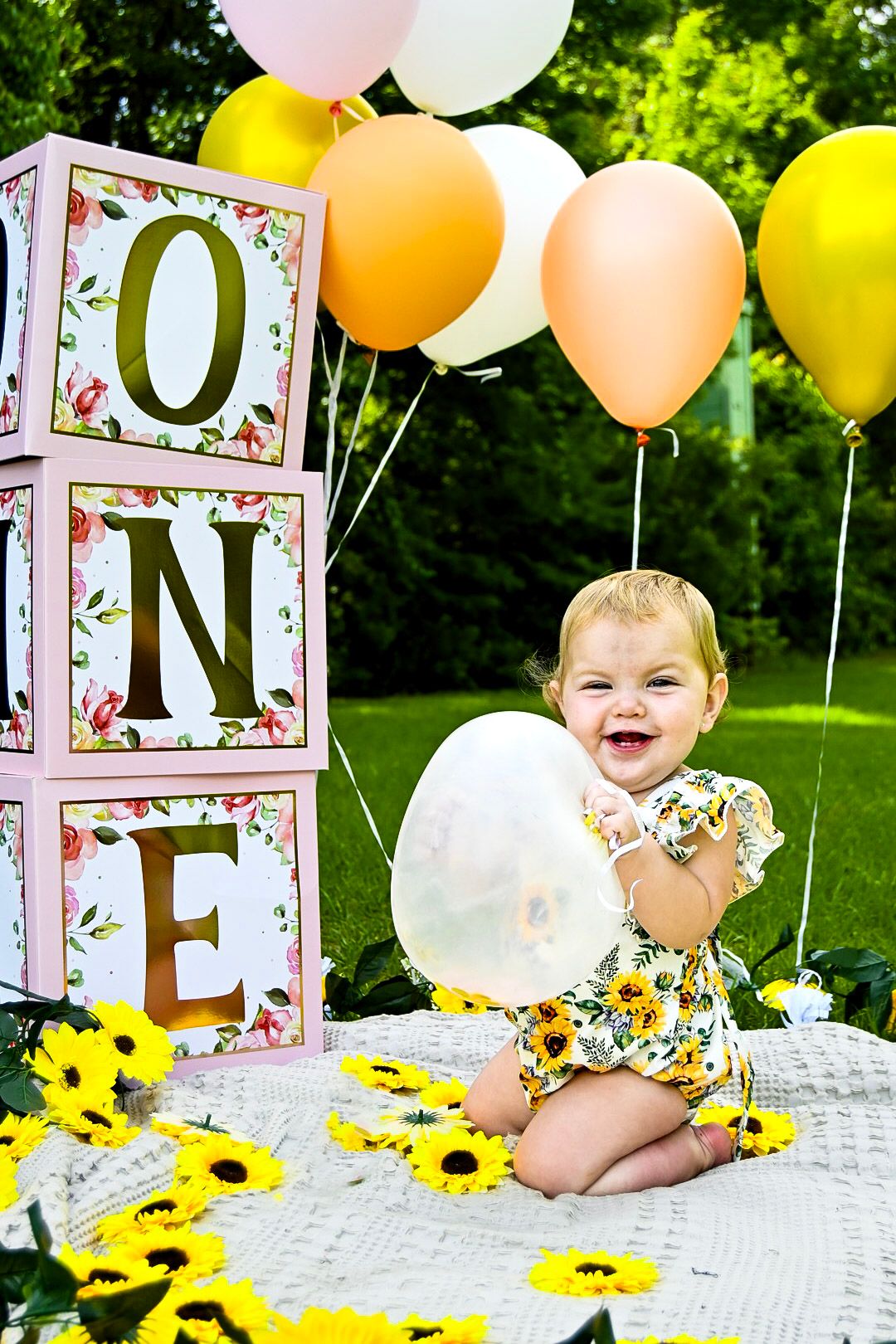 Baby sits on blanket with decorative letter blocks and sunflowers for a first birthday photoshoot.