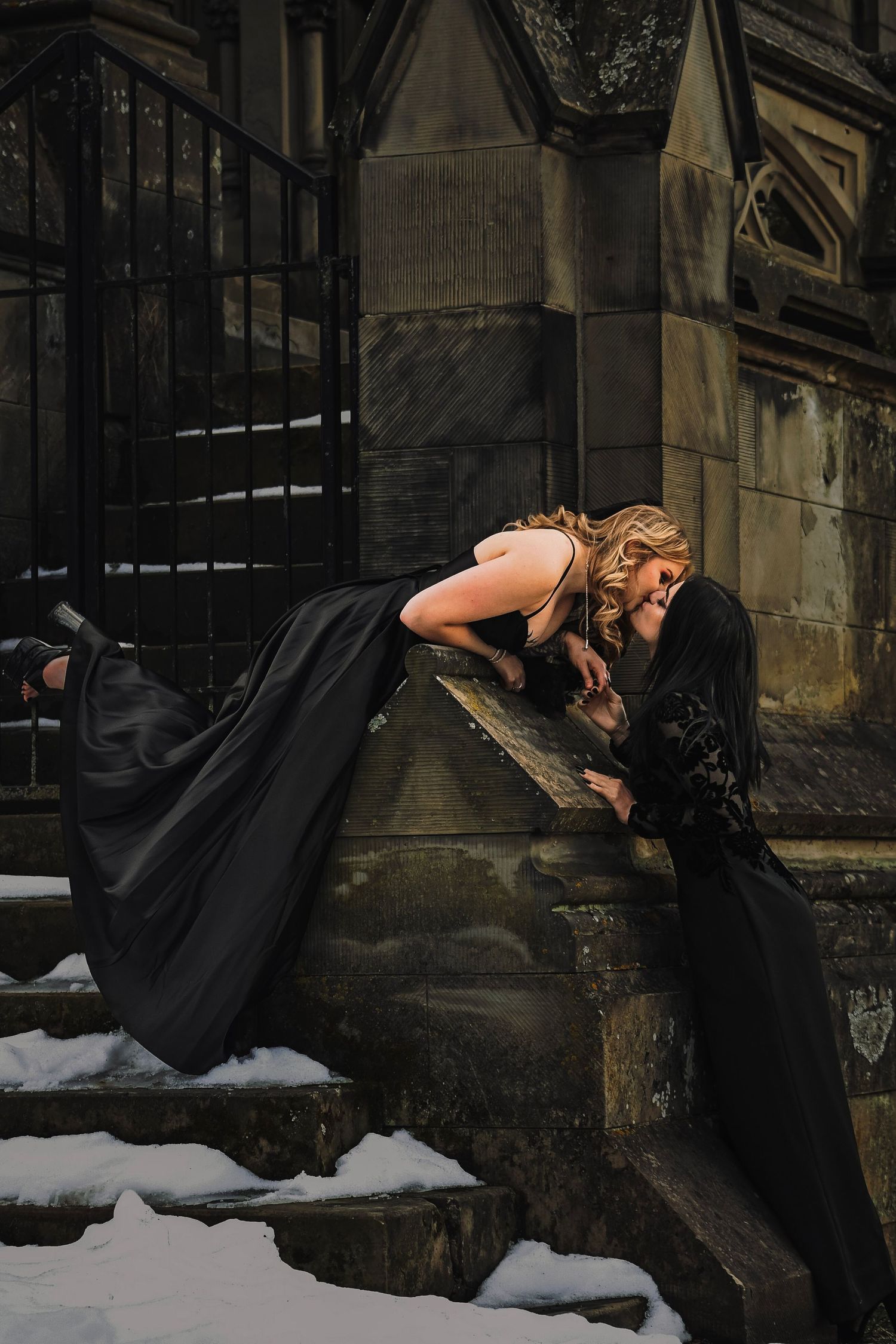 A person in black clothing bends over a snowy surface near a dark stone building in dramatic lighting.