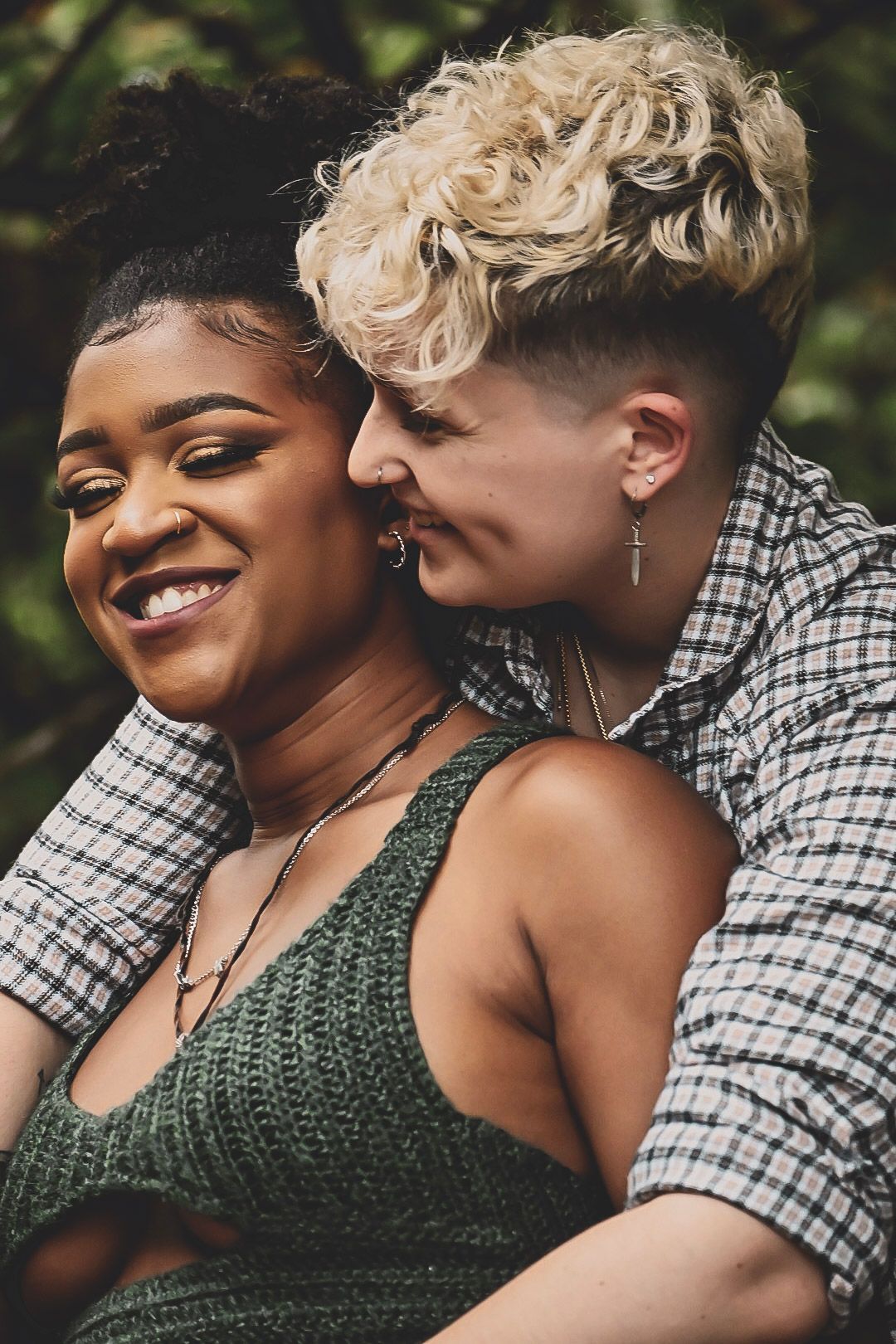 Two women sharing a tender moment outdoors, one smiling brightly as the other leans in affectionately.