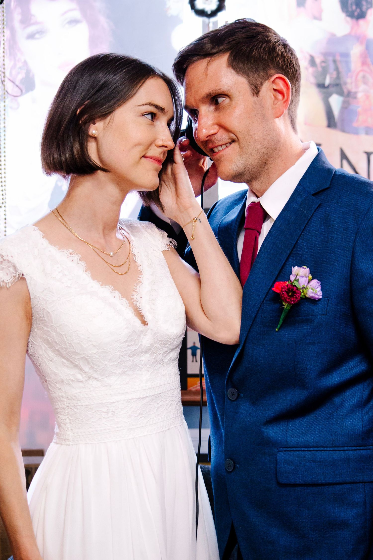 A bride in white lace dress and groom in blue suit with red tie share a tender moment at their wedding ceremony.
