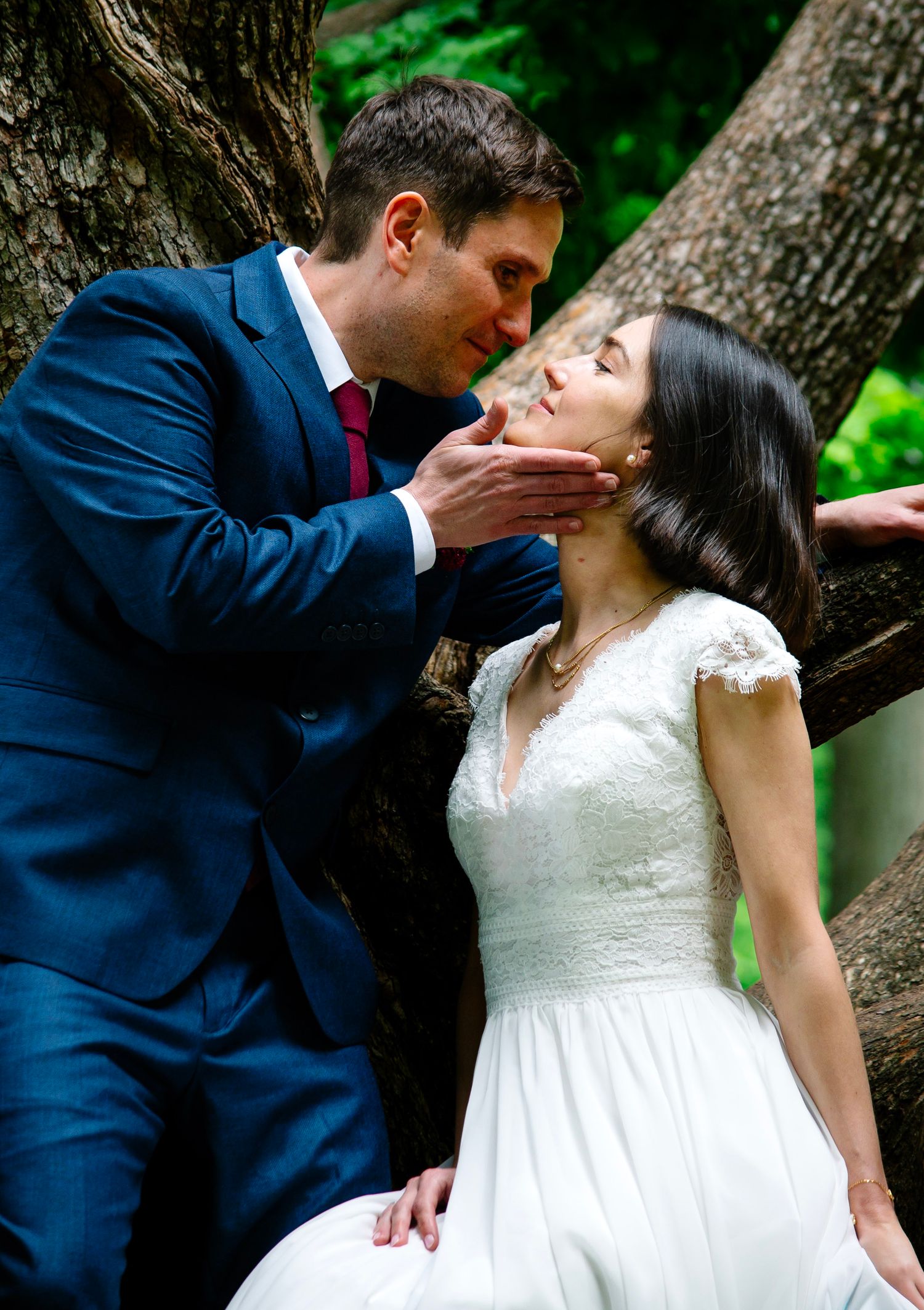 Groom in blue suit tenderly cups bride's face for a kiss outdoors near trees in a romantic wedding portrait.