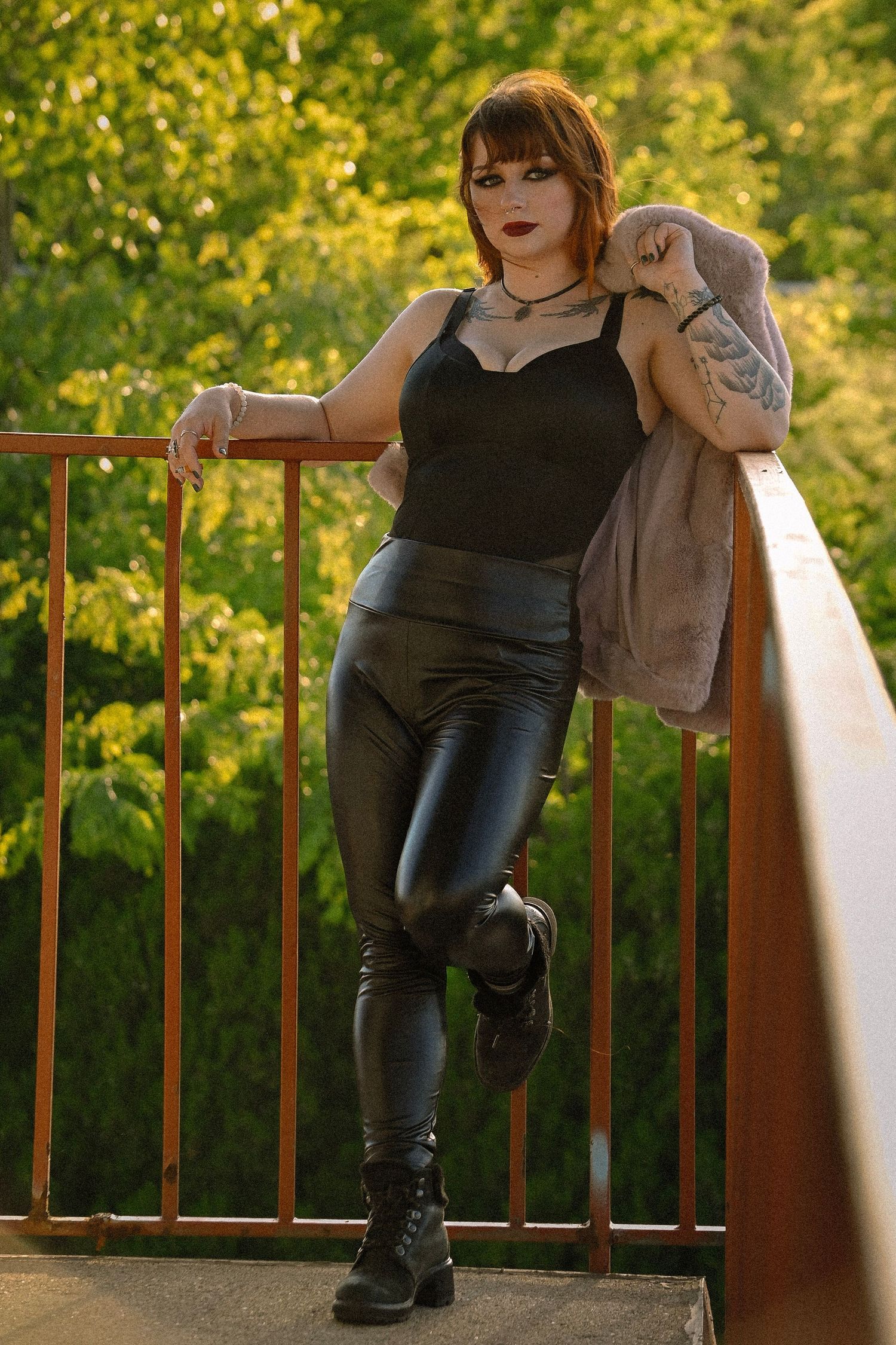 Woman in black bodysuit, leather pants, and boots posing on a bridge railing with green trees behind her.