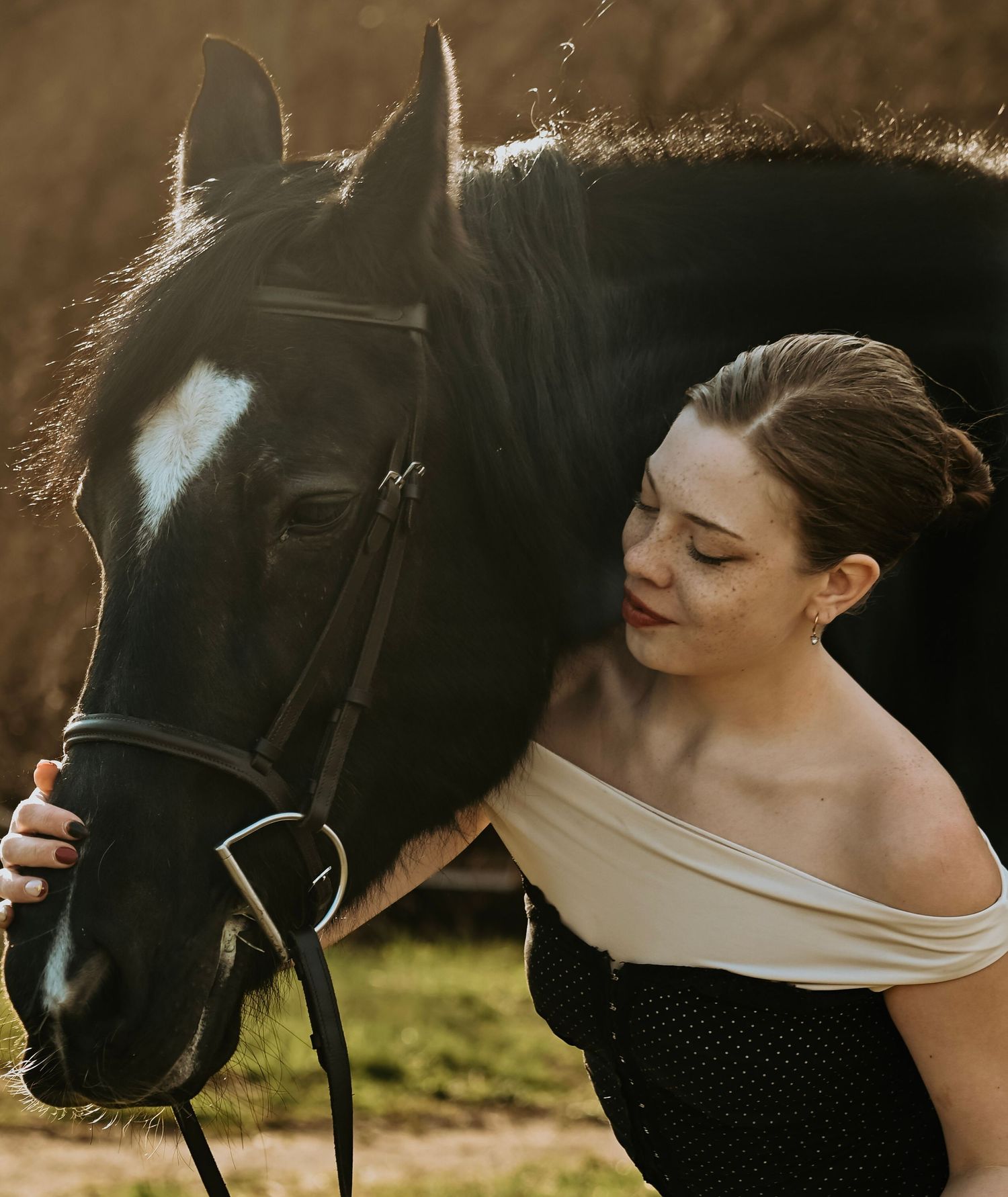 Woman in black dress poses with a black horse in a scenic outdoor setting during golden hour lighting.