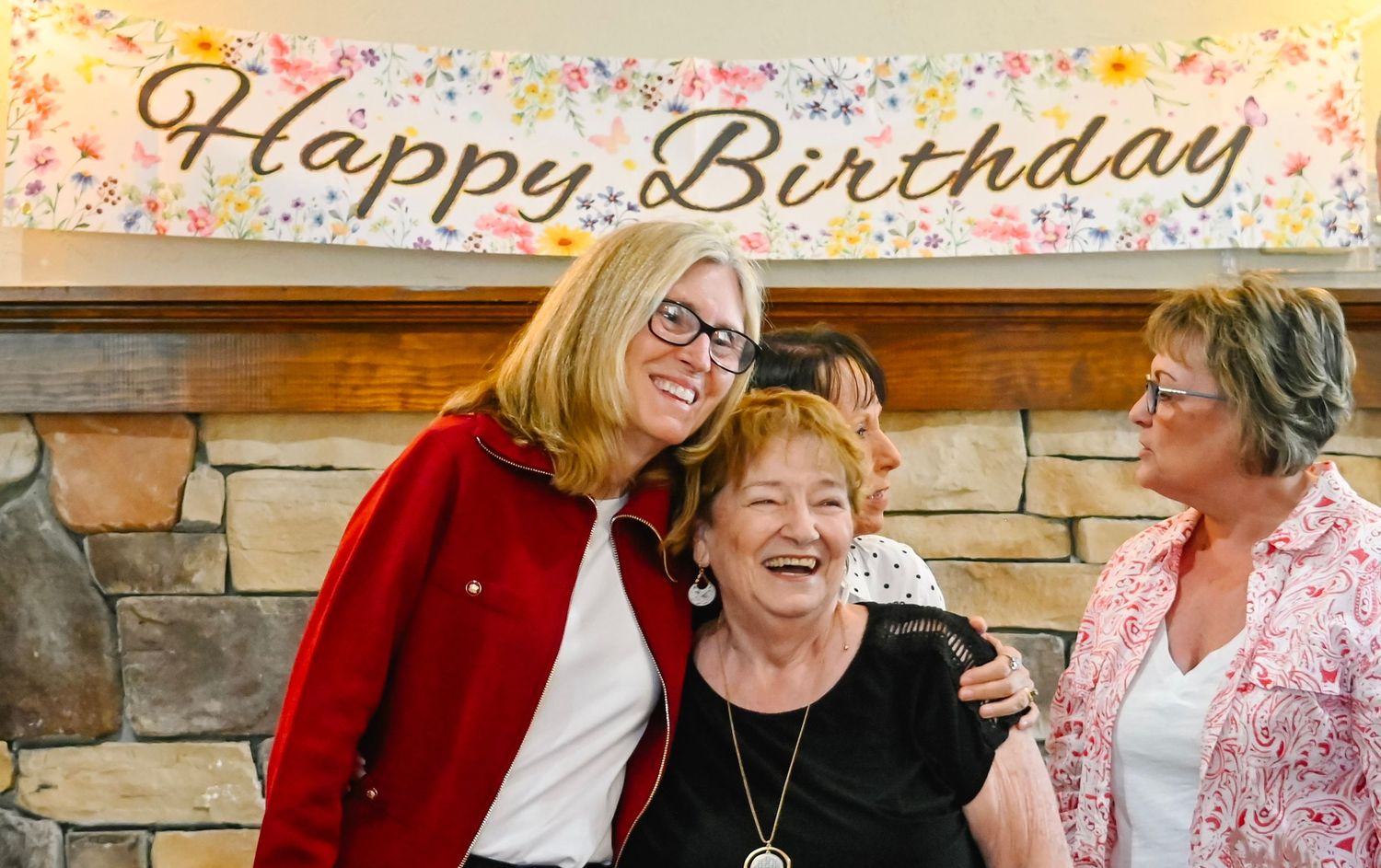 Three women smile together under a Happy Birthday banner at a birthday celebration party.