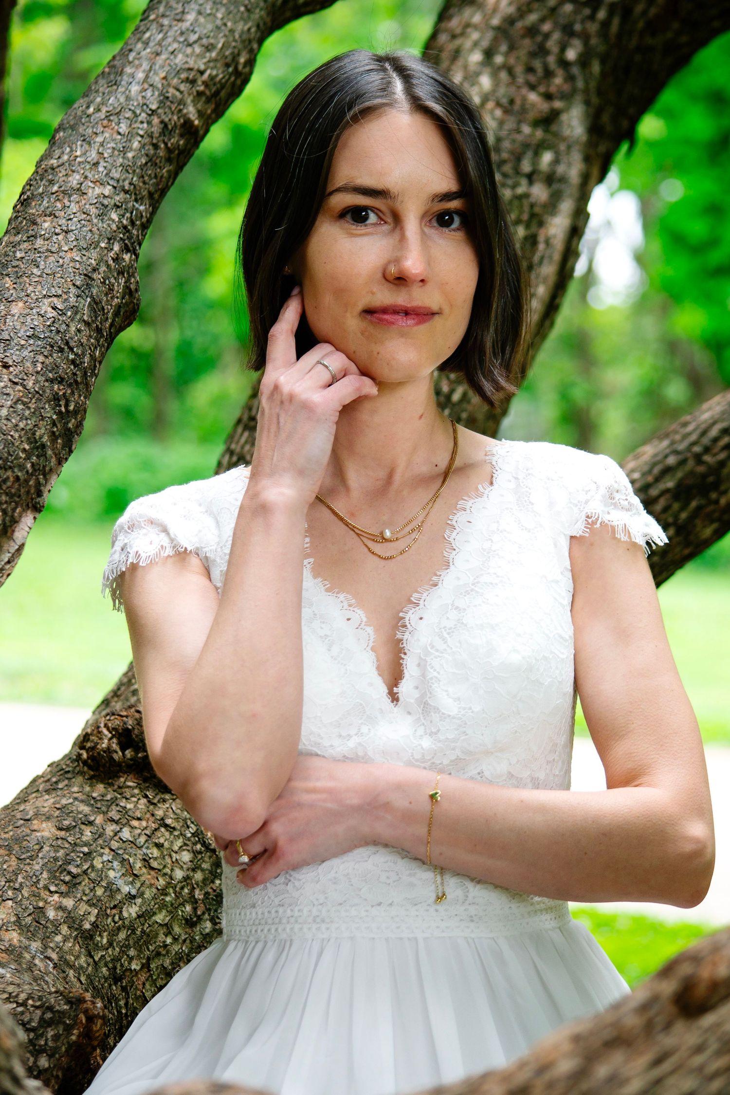 Elegant bride in white lace dress poses gracefully against a tree in a lush green park setting.