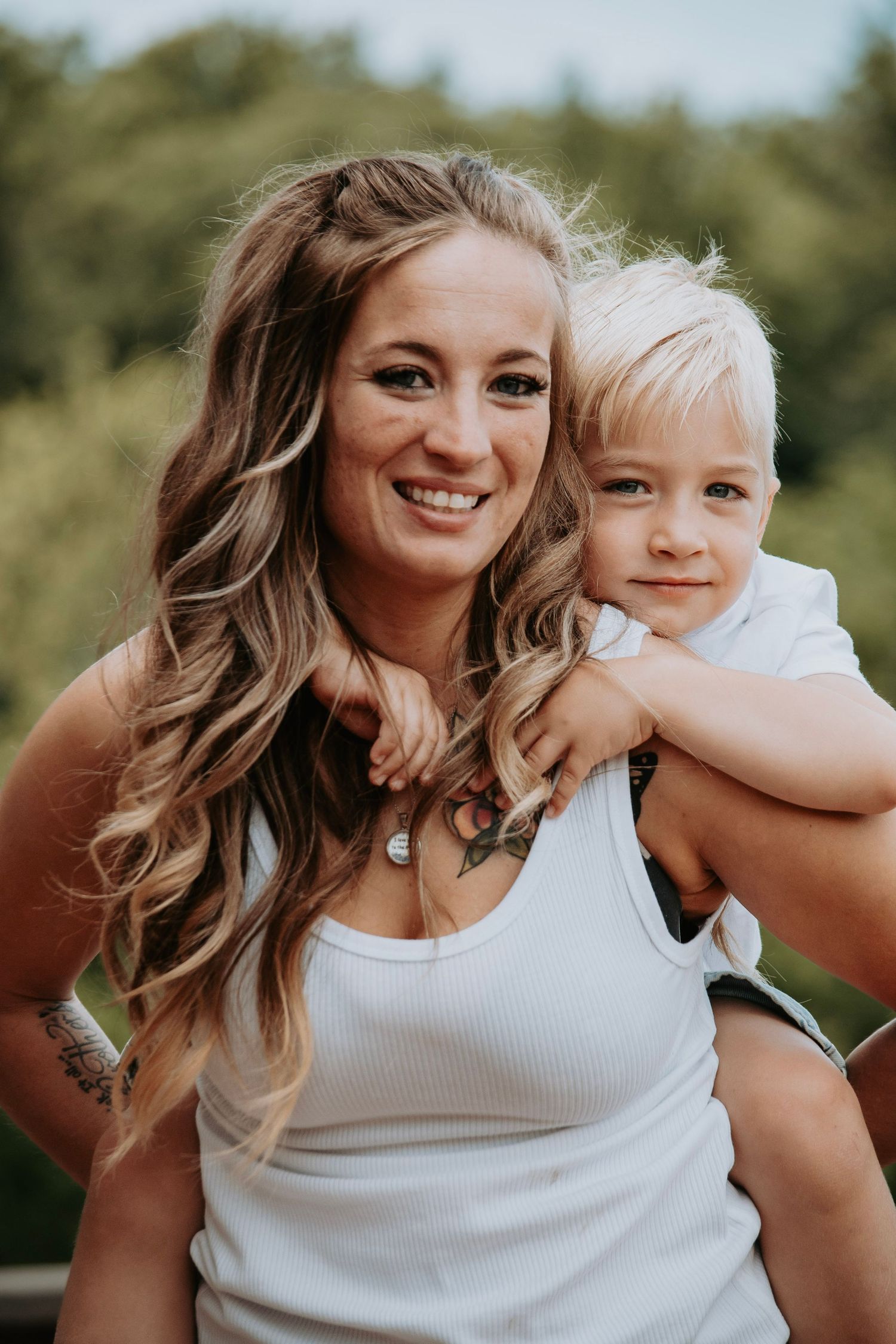 Woman with long wavy hair smiling while holding a young blonde boy on her back outdoors.