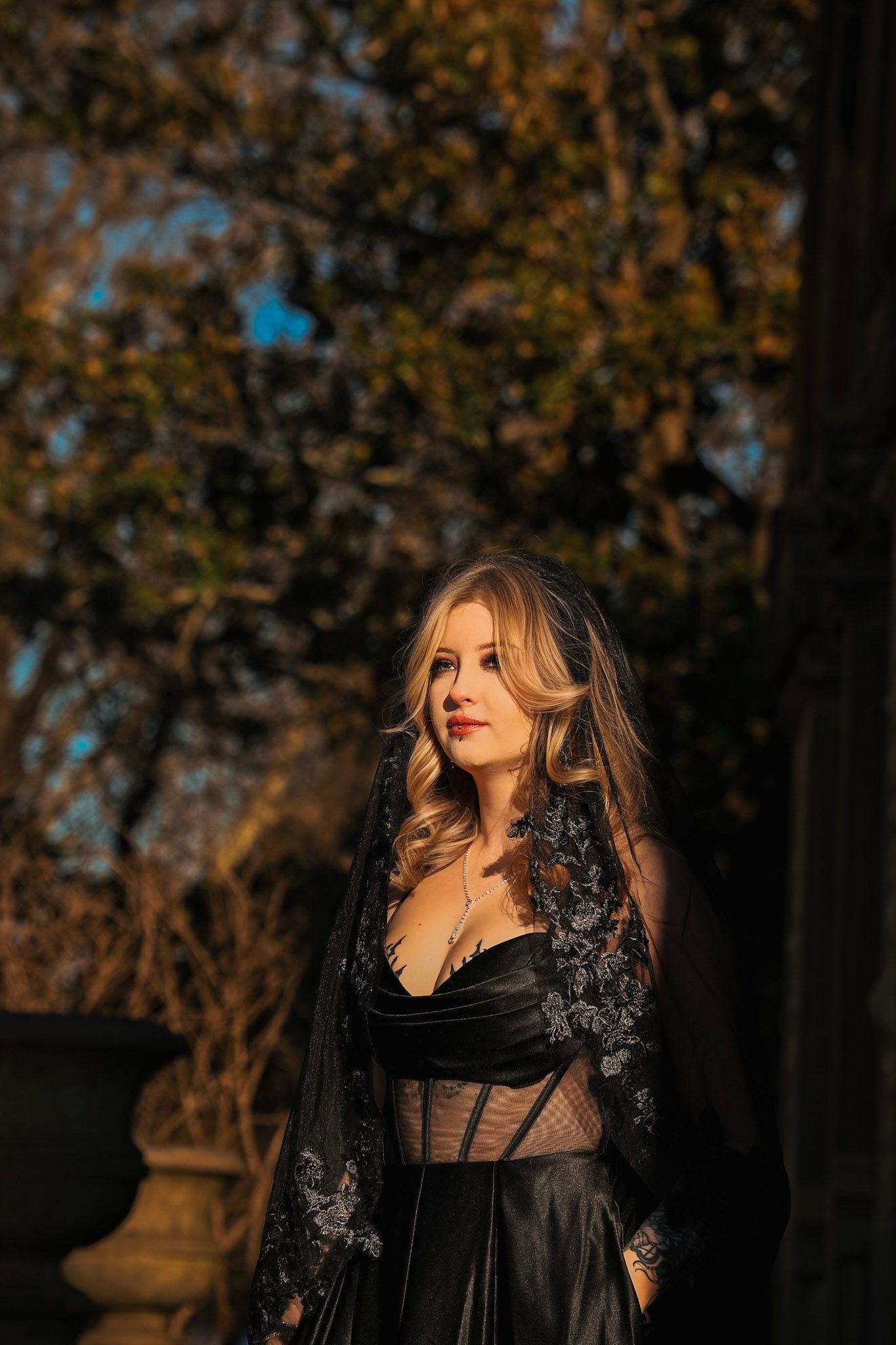 A woman in a black gothic dress poses dramatically in an outdoor setting with autumn trees in the background.