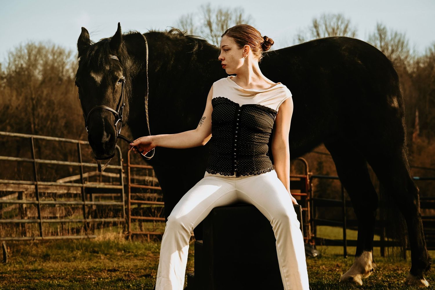 Woman in black corset and white pants poses with a black horse at a rustic fence outdoors.
