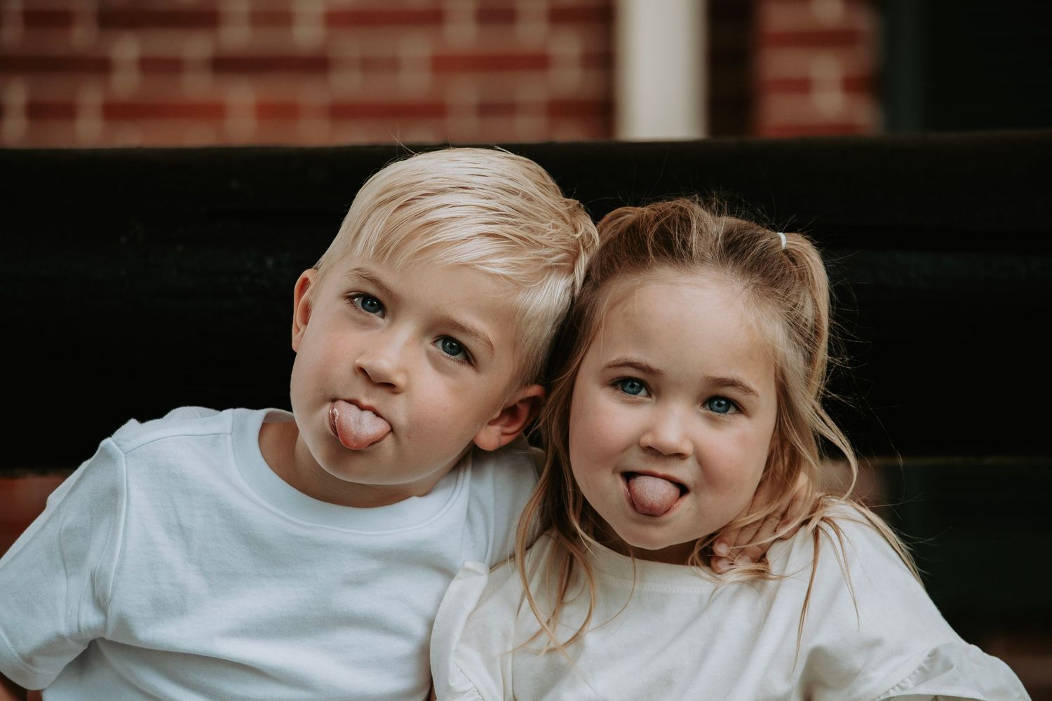 Two young children in white shirts sit together on steps, playfully sticking out their tongues.