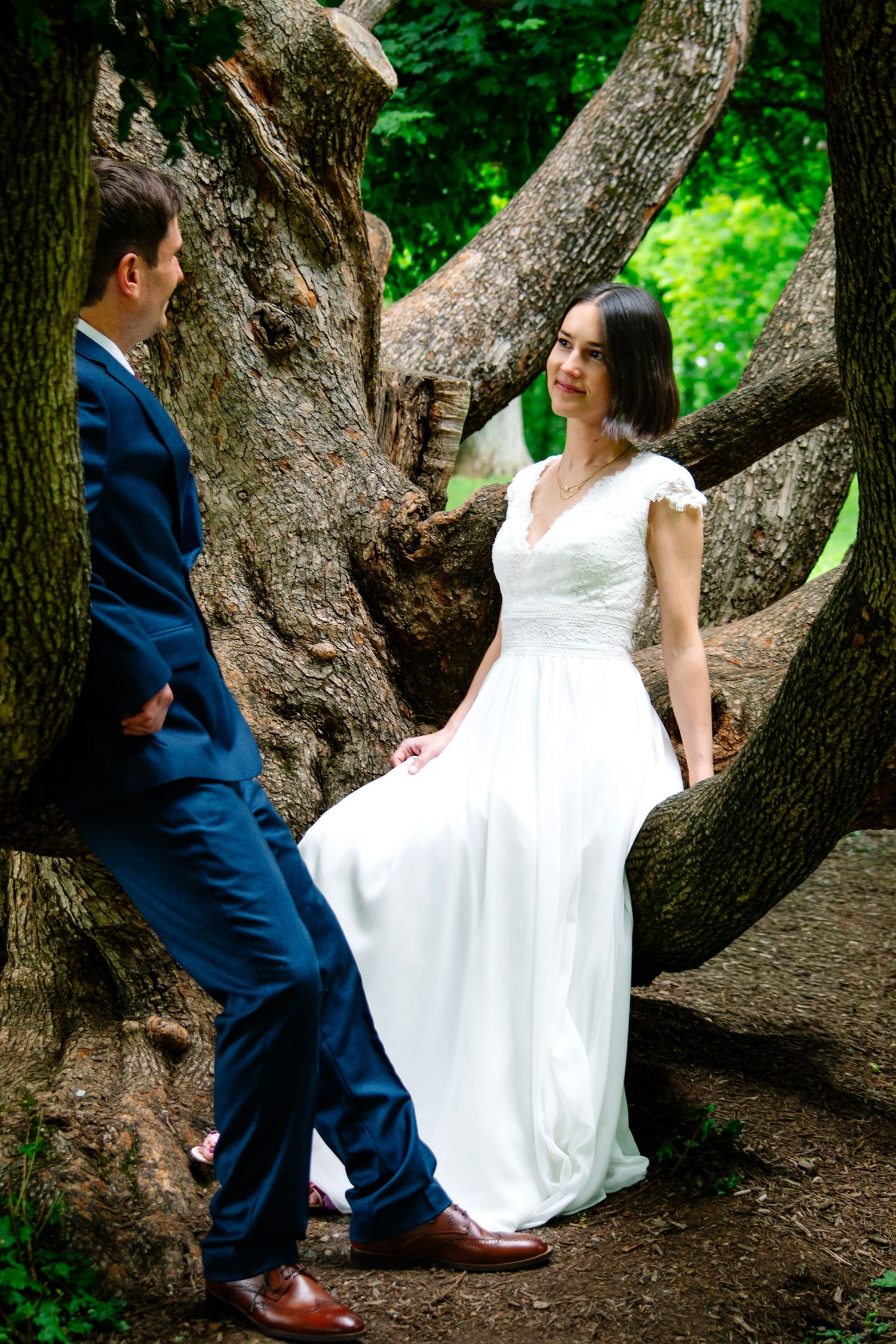 Bride in white gown sits on large tree roots as groom in navy suit stands beside her in lush green park.