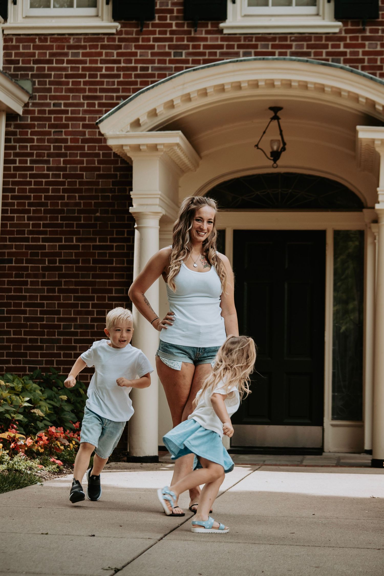 A smiling woman watches two young children playing outside a brick house with white columns.