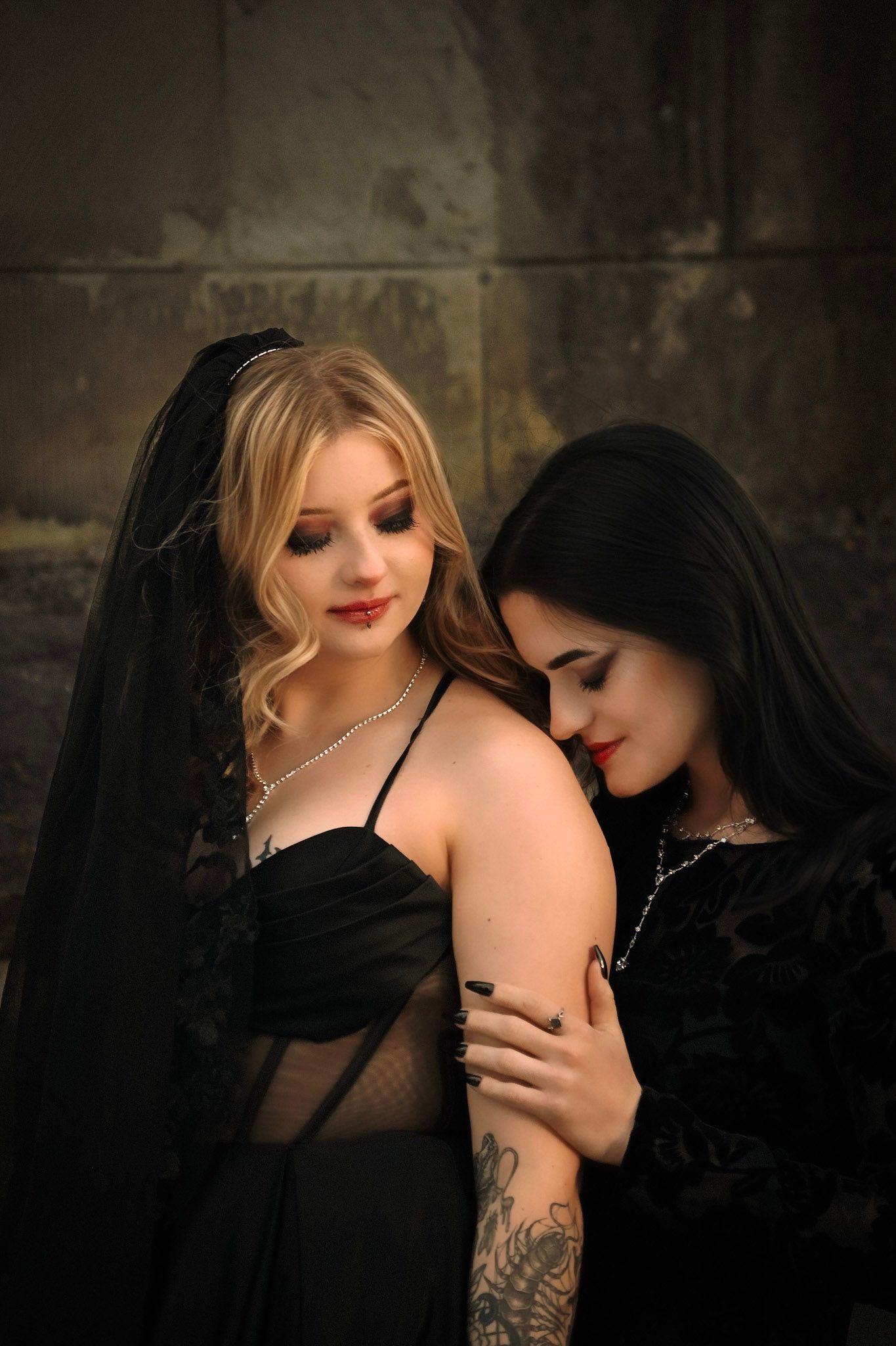 Two women in black gothic dresses and veils pose intimately against dark textured wall with moody lighting.