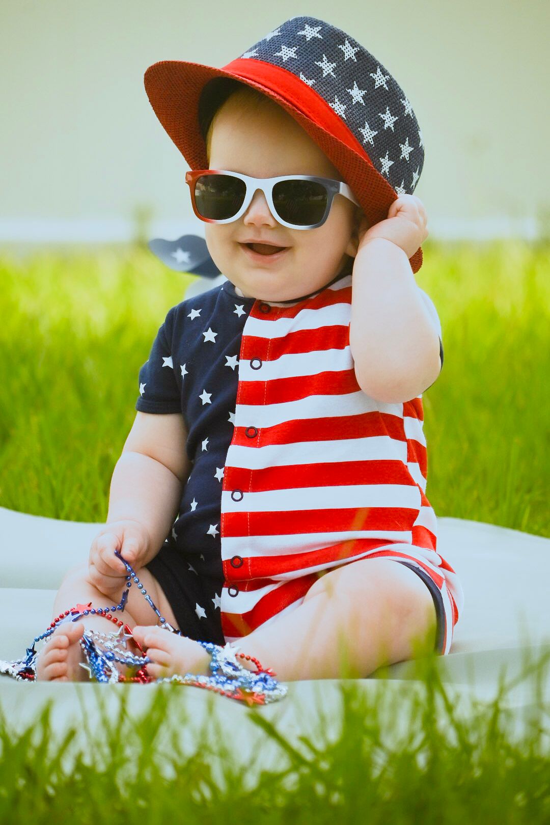 Baby wearing American flag outfit and sunglasses sitting on blanket outdoors.