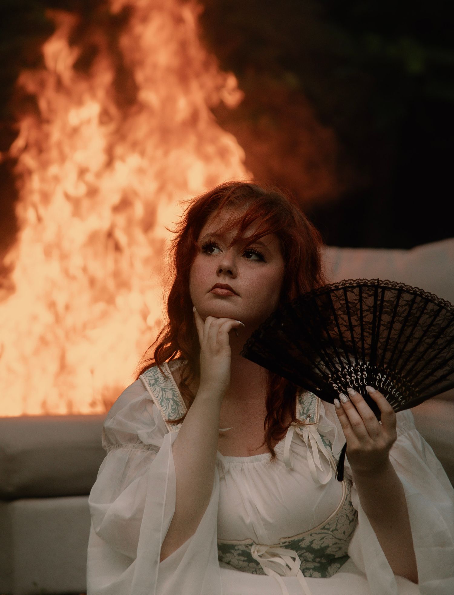 Red-haired woman in white dress holds black fan, posing dramatically before large flames.