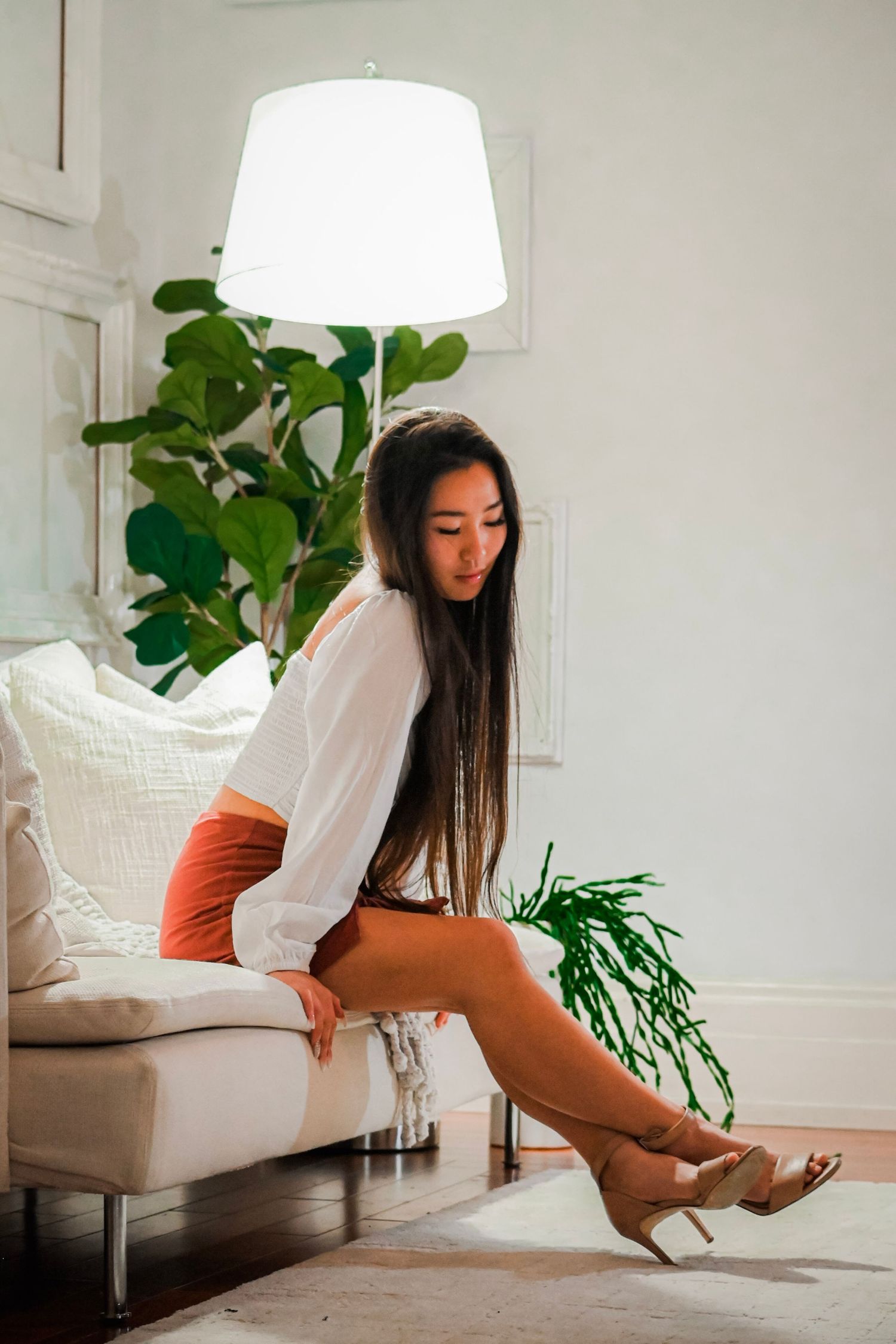 Woman in white top and orange shorts sits elegantly on white couch beneath modern lamp fixture.