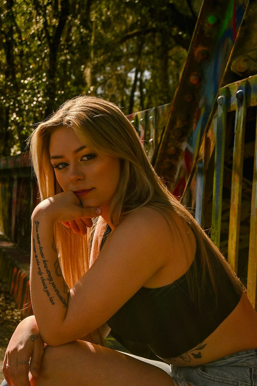Person in black tank top posing outdoors during golden hour with tattoos visible.