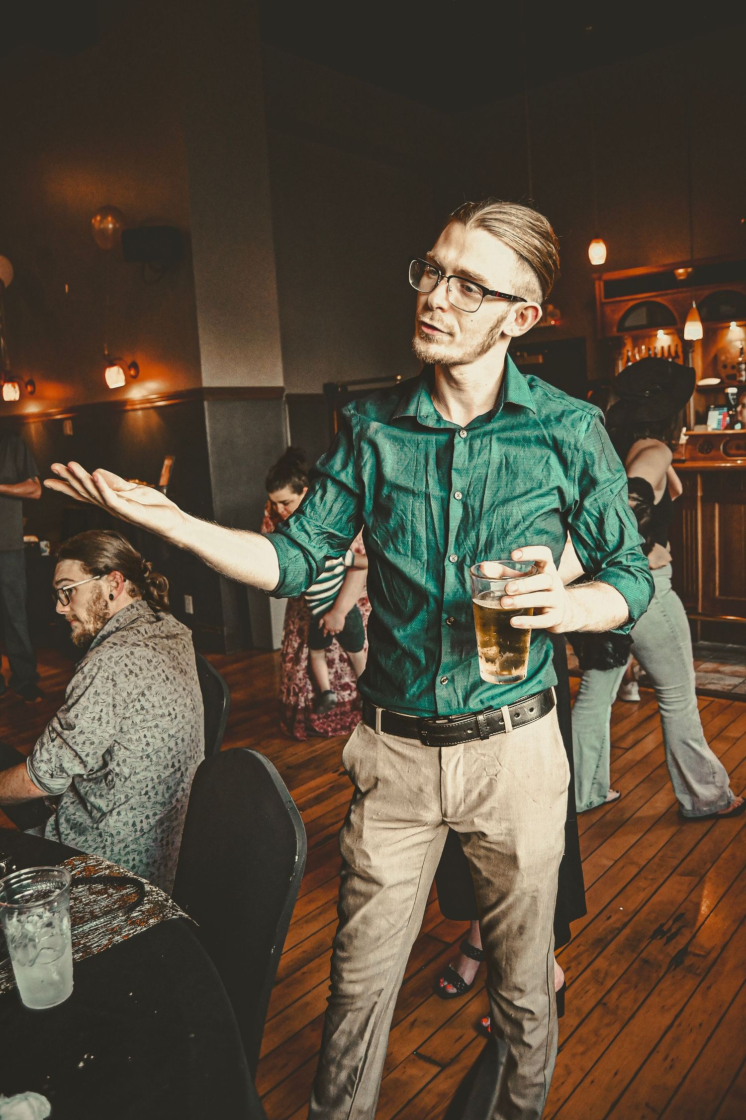 A person in a green shirt gesturing expressively while holding a drink at an indoor gathering.