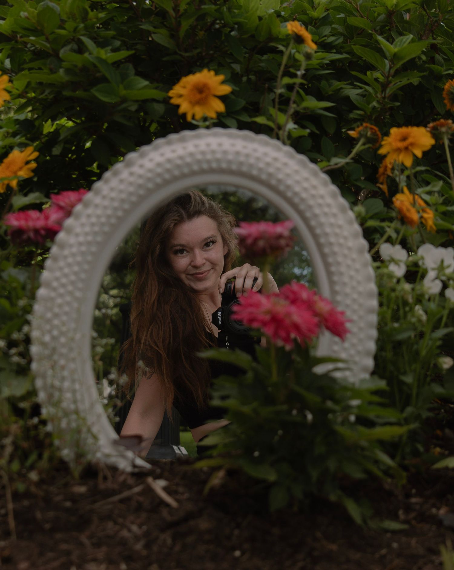 Young woman smiling through a white decorative arch surrounded by colorful flowers in a lush garden.