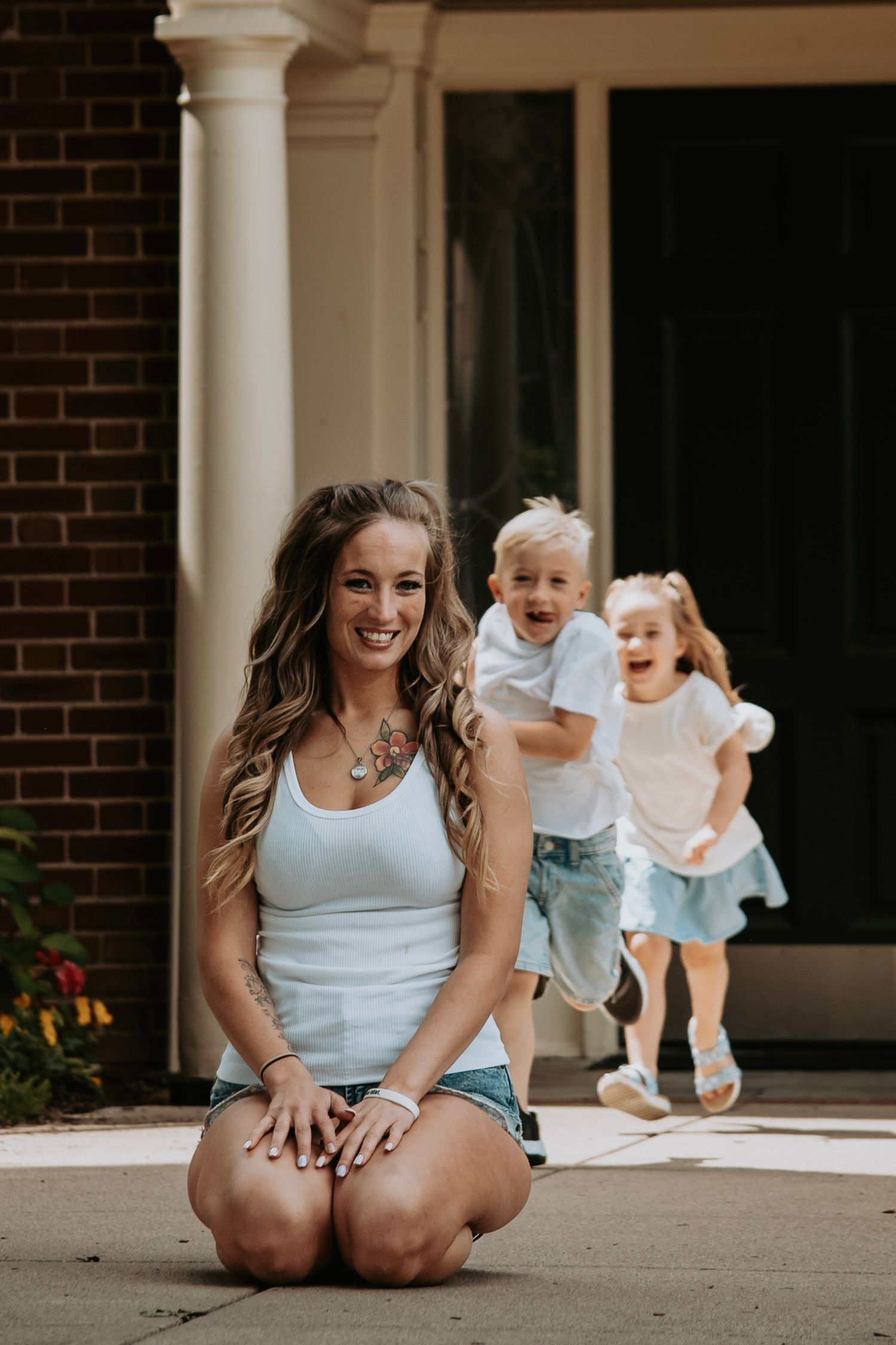 A smiling woman with long hair sits on a driveway as two young children run toward her in front of a brick home.