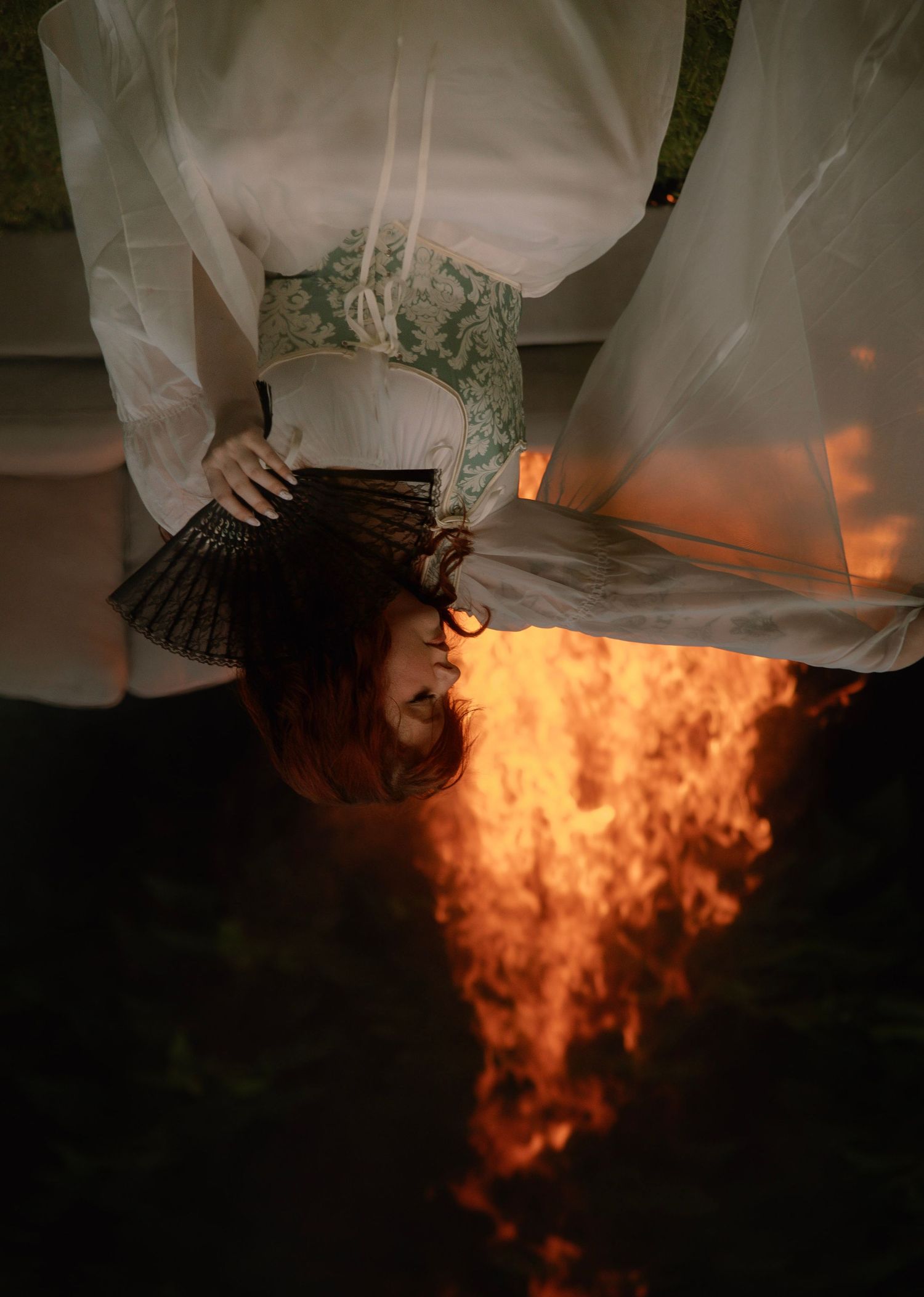 A red-haired woman in a white corset dress photographed upside down above a blazing fire outdoors.