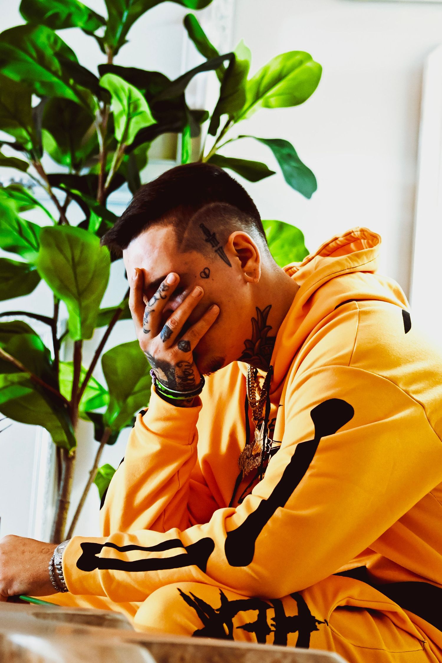 Person in bright orange hoodie sitting thoughtfully with hand on face next to large indoor plant.