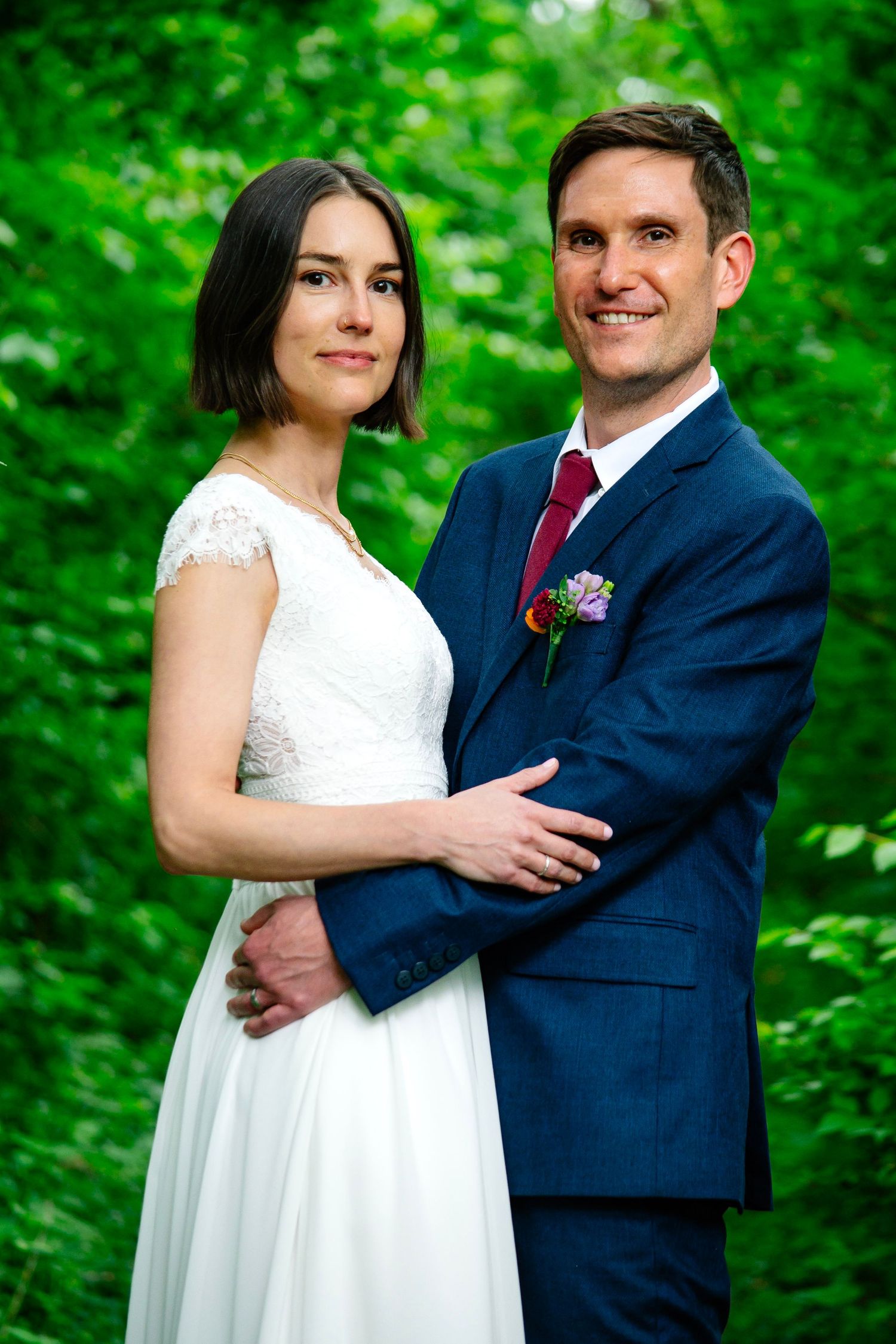 Bride in white lace dress and groom in navy suit with boutonniere pose together outdoors among lush green foliage.