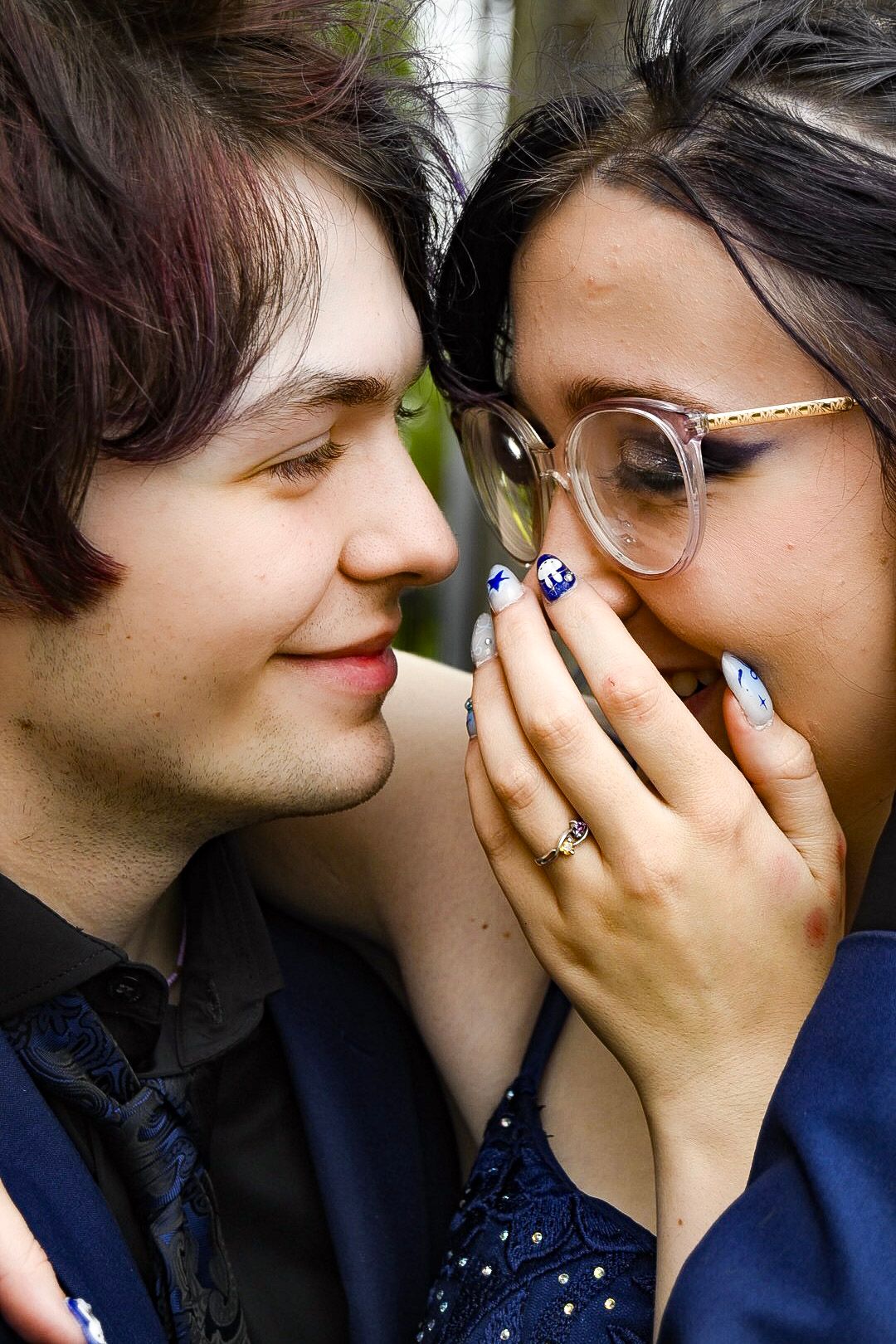 A young couple shares an intimate moment, the woman laughing behind decorated blue nails while wearing glasses and a blue dress.