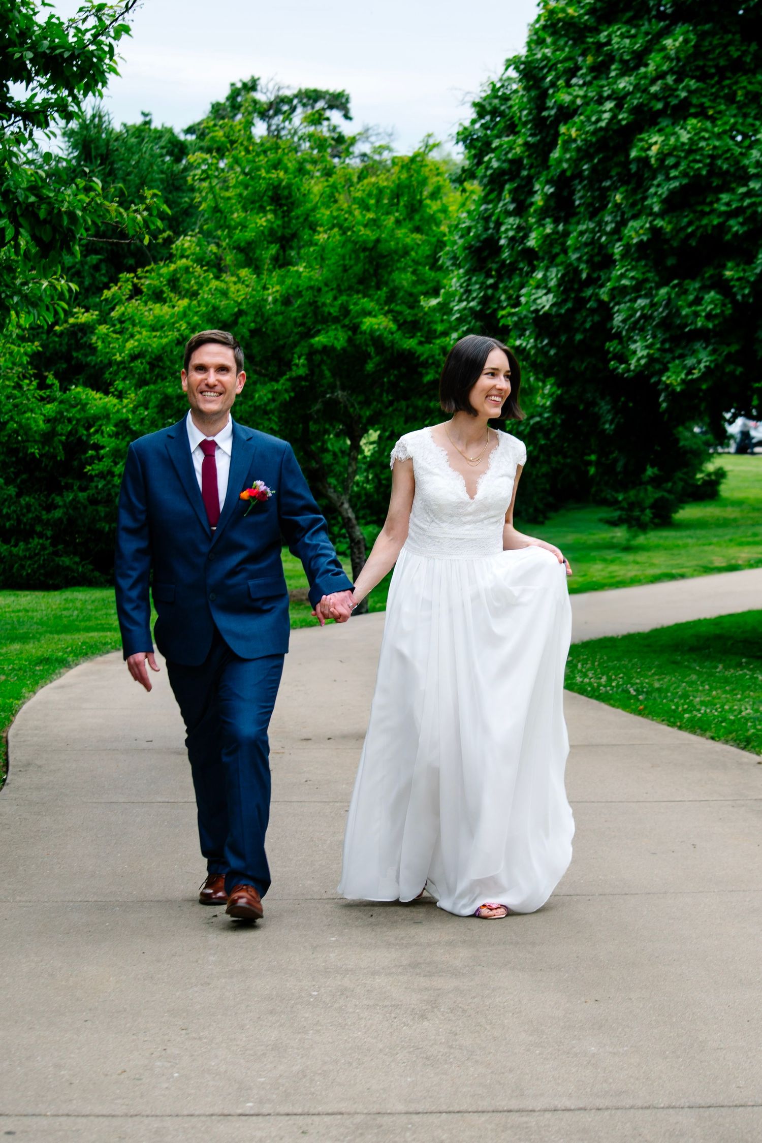 A smiling bride and groom walk hand-in-hand on a park path, she in a white gown, he in a navy suit with red tie.
