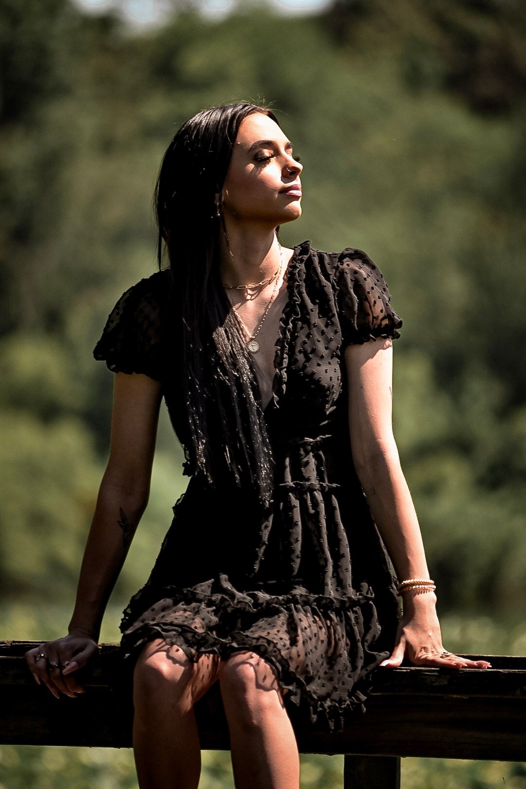 Person in black lace dress sitting on wooden railing in natural setting.