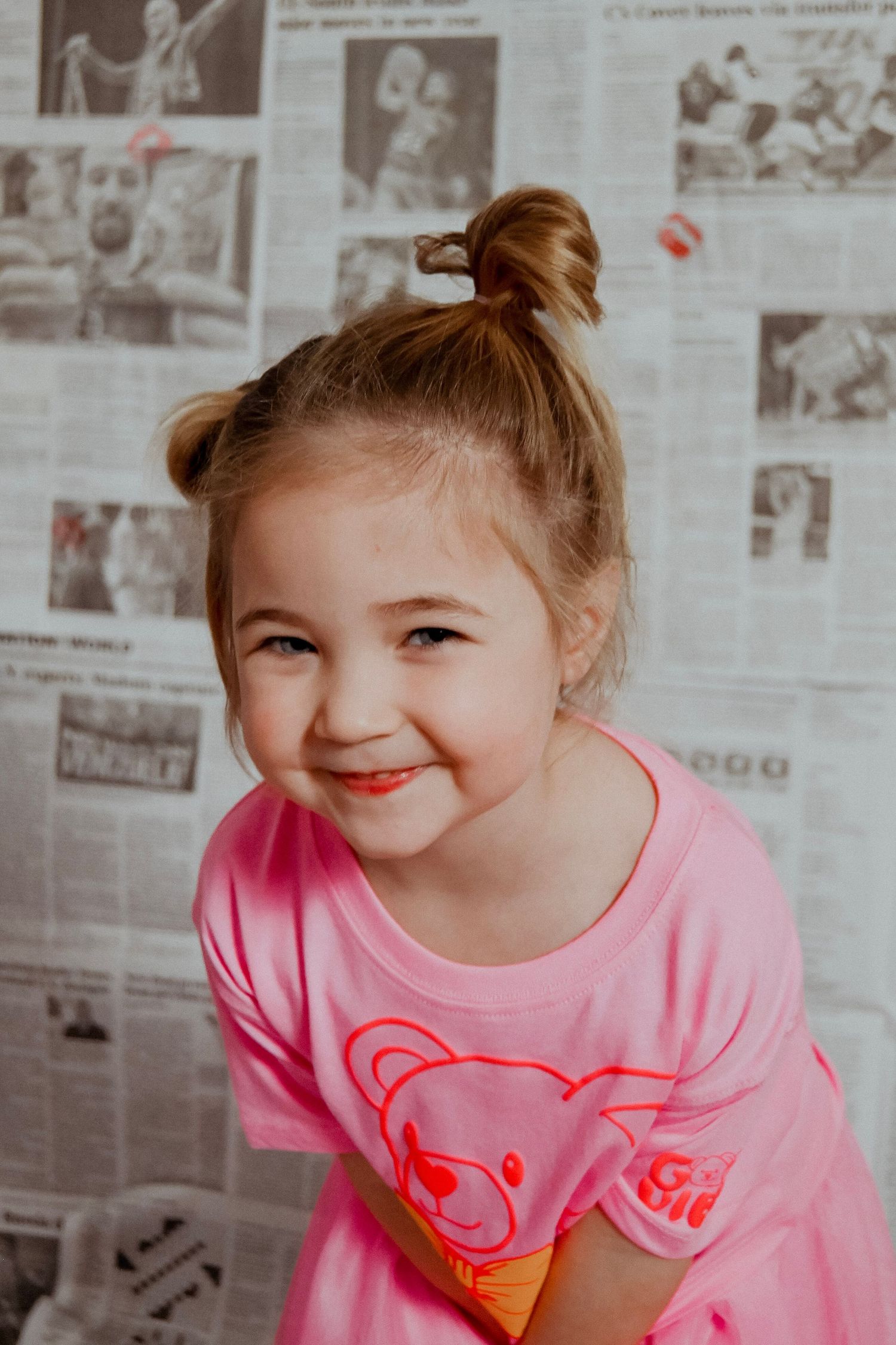 Young child wearing pink shirt smiling against newspaper backdrop with red heart decorations.