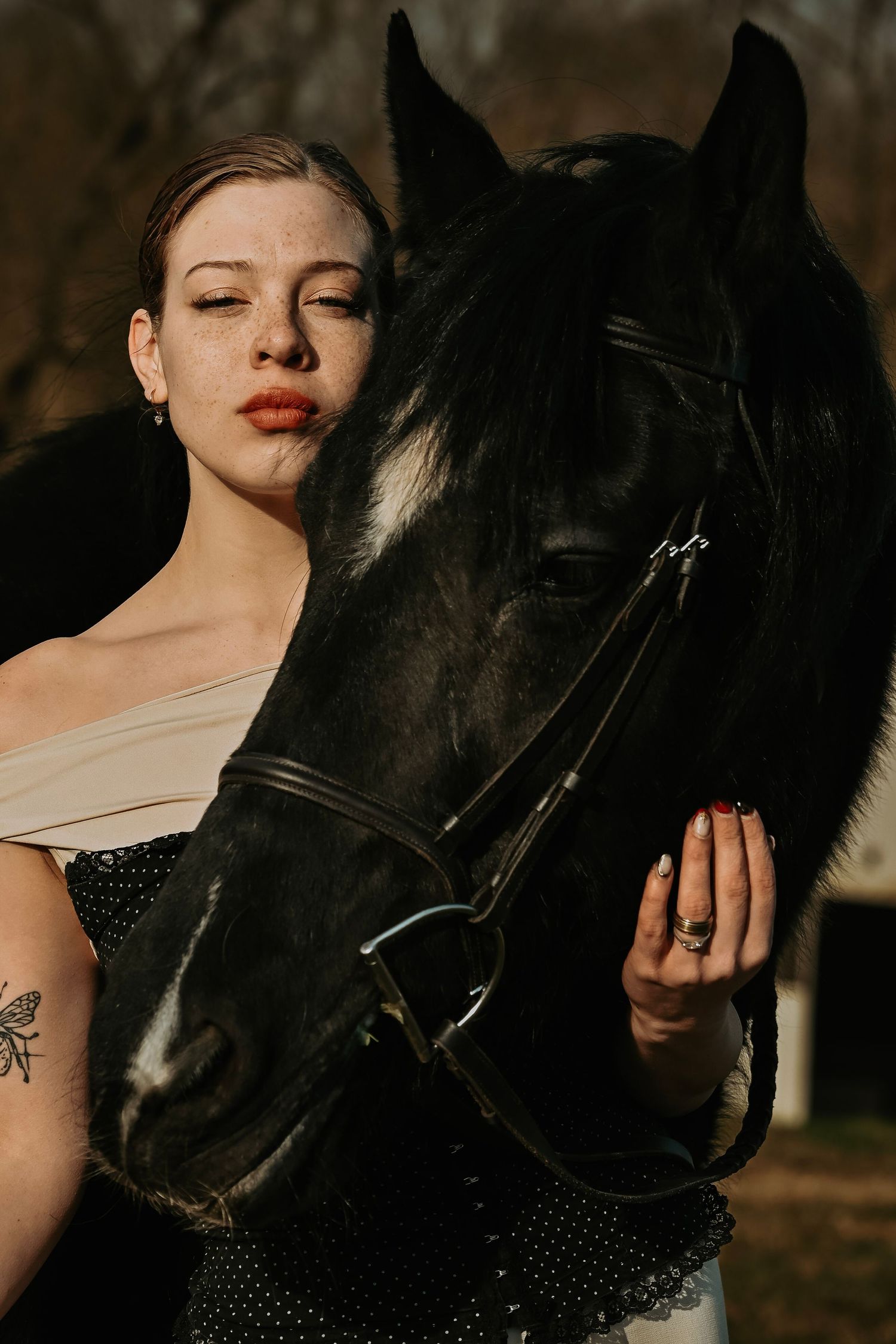 Woman with red lips and tattoo posing closely with a black horse wearing a bridle outdoors.