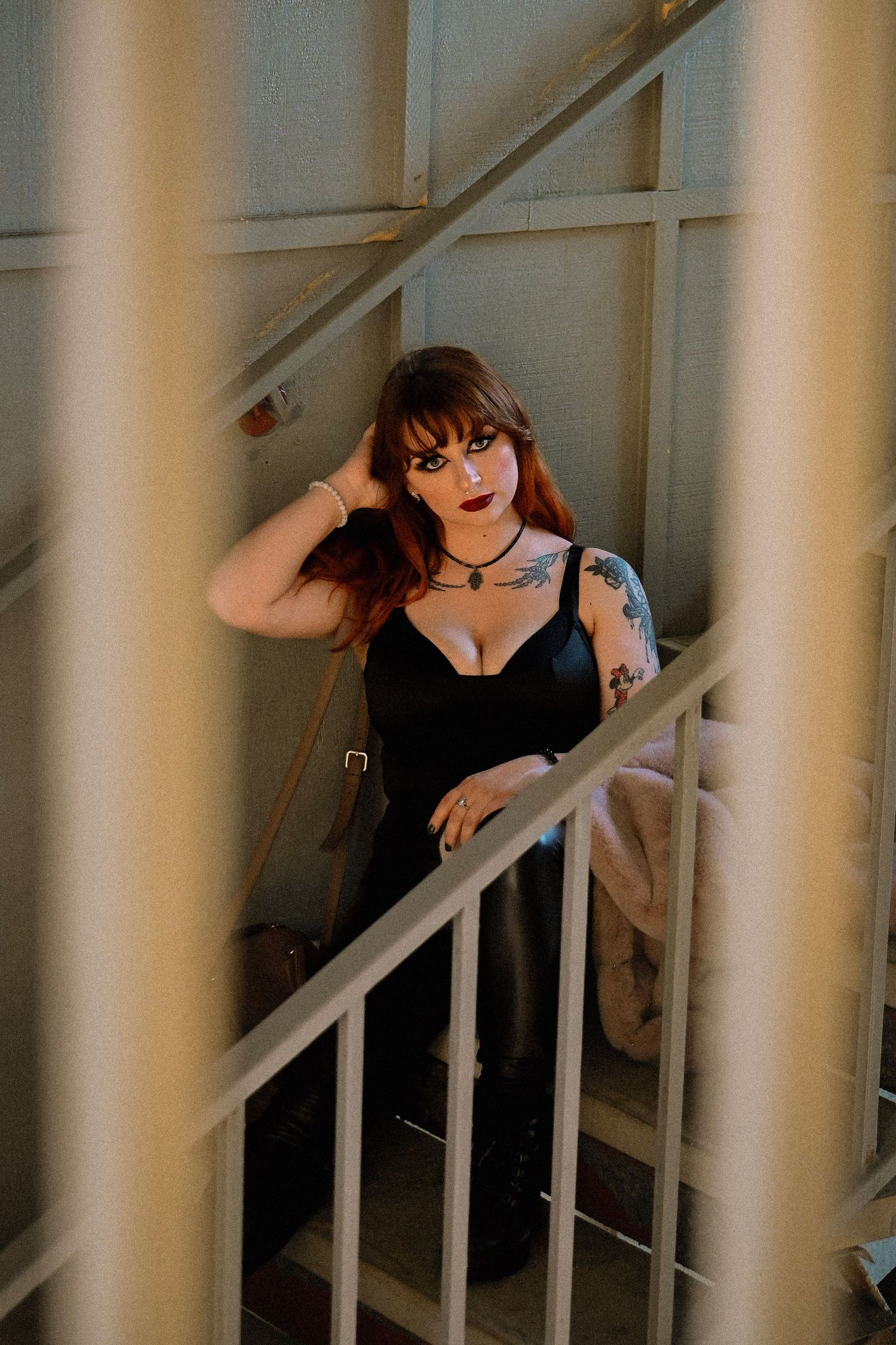 A tattooed redhead woman in a black outfit poses on a staircase with gothic makeup and a choker necklace.
