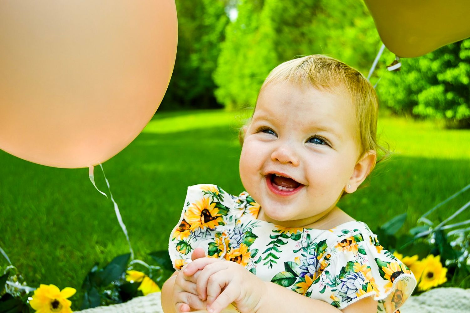 Baby in a floral outfit plays with colorful balloons on a sunny day.
