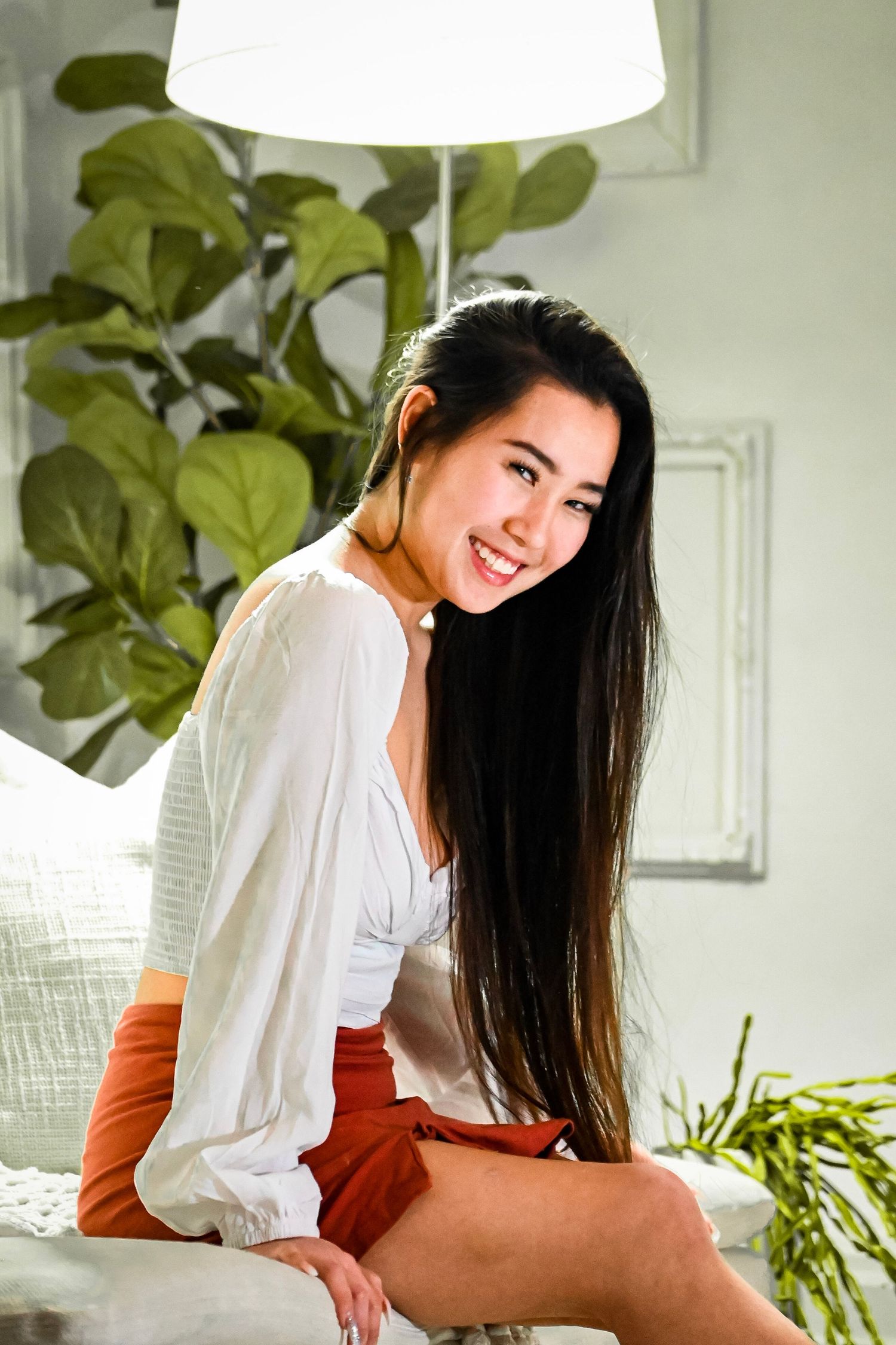 Woman in white blouse smiling while working at desk with plants in bright room.