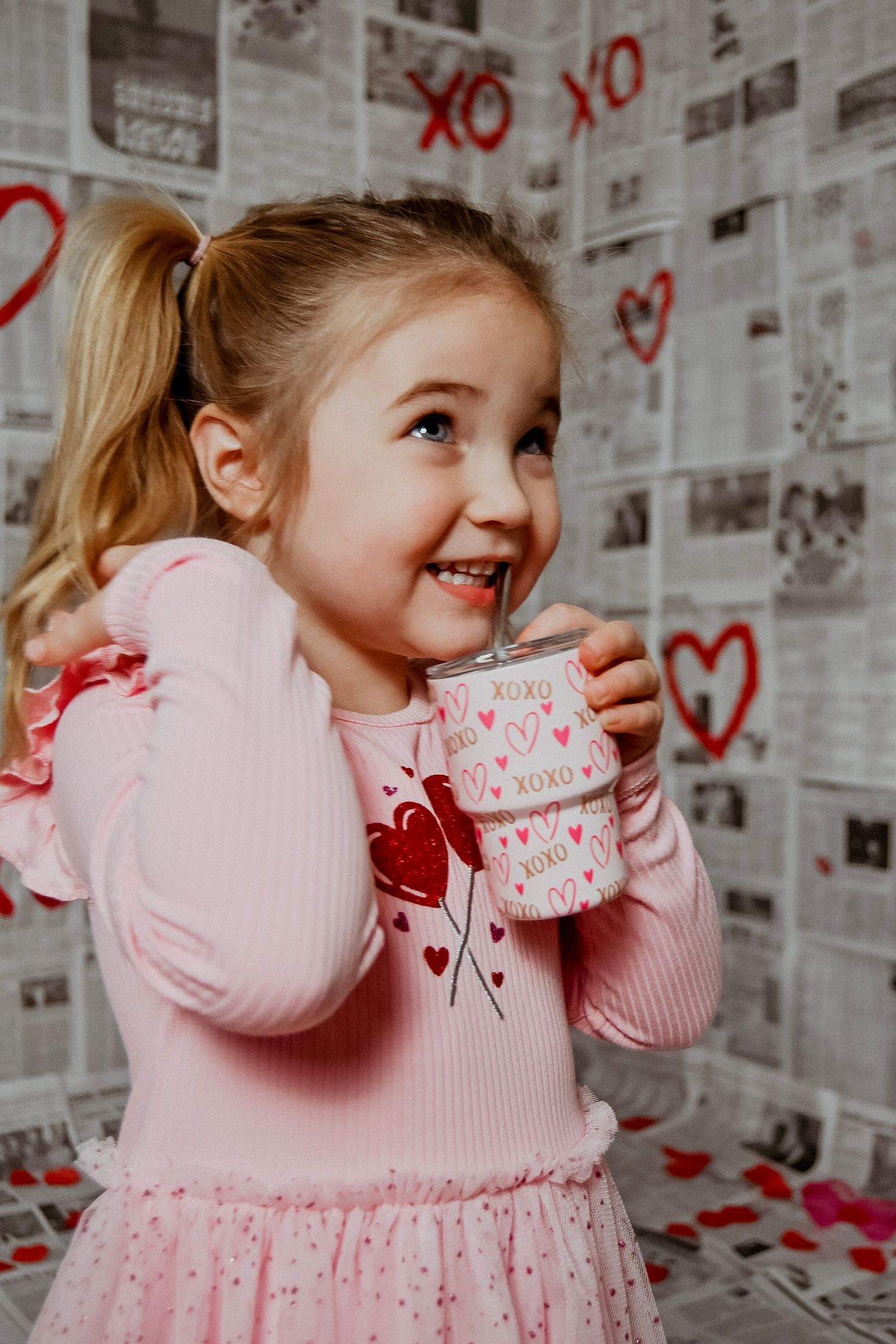 Young child in pink sweater holding Valentine's mug against wall decorated with hearts and XO letters.